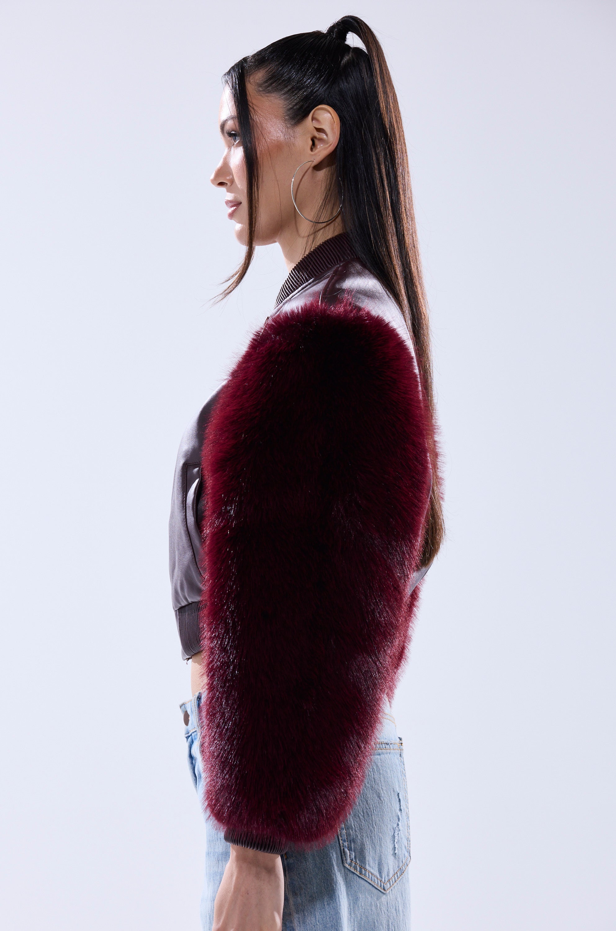 A woman with long dark hair in a ponytail models the SO GOOD IT HURTS FUR SLEEVE BOMBER IN BURGUNDY, pairing it with hoop earrings and light blue jeans, standing in profile against a plain light background.
