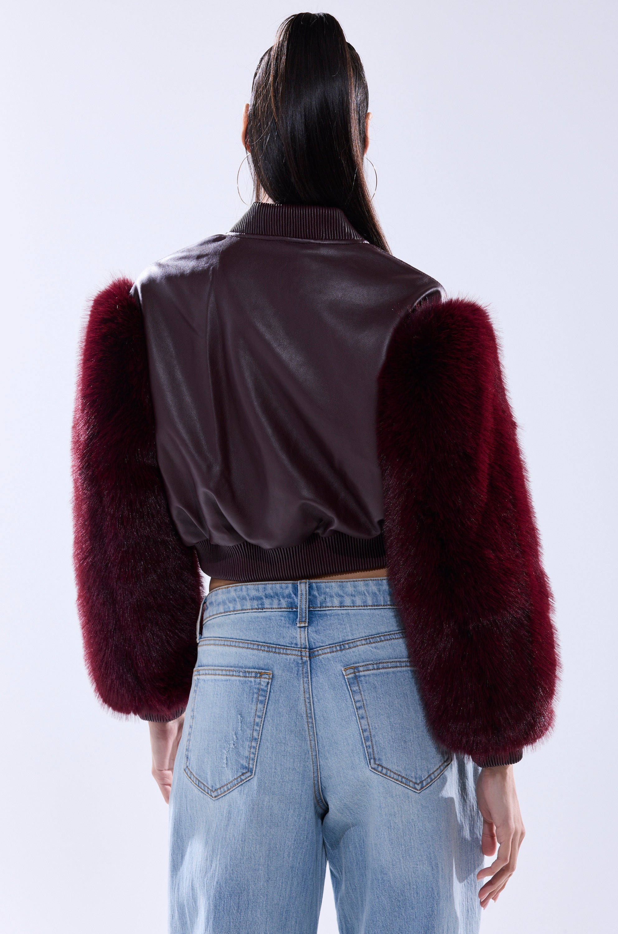 Wearing the SO GOOD IT HURTS FUR SLEEVE BOMBER IN BURGUNDY with voluminous dark red faux fur sleeves and light blue jeans, a person is seen from the back against a plain light background.