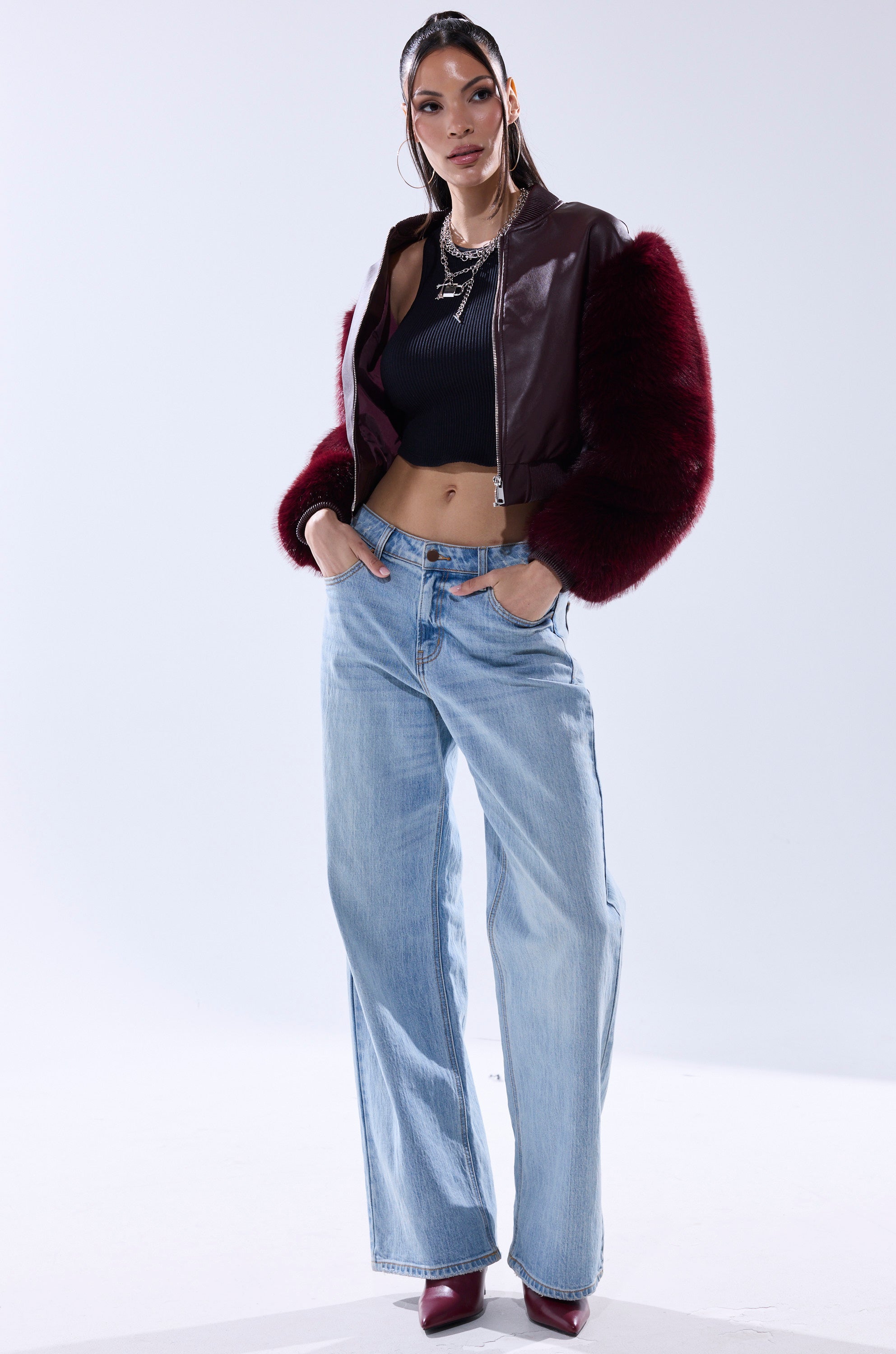 A woman stands confidently in the SO GOOD IT HURTS FUR SLEEVE BOMBER IN BURGUNDY, styled with a black ribbed crop top, light blue wide-leg jeans, maroon pointed-toe heels, long hair, hoop earrings, and layered necklaces.