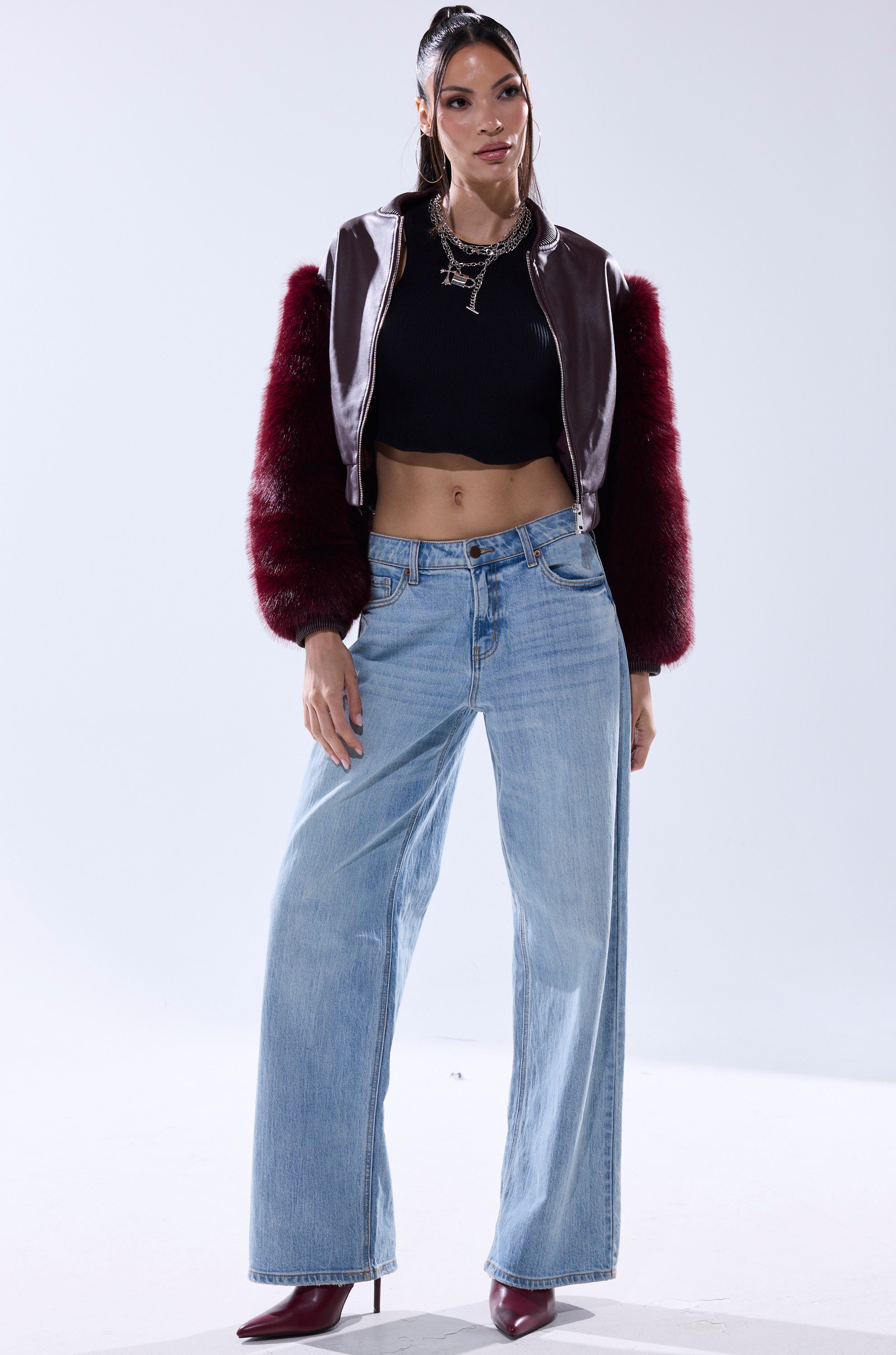 A woman stands confidently in a black cropped top, SO GOOD IT HURTS FUR SLEEVE BOMBER IN BURGUNDY, light blue wide-leg jeans, and maroon heeled boots. Her long dark hair is tied back and she wears layered necklaces against a plain background.