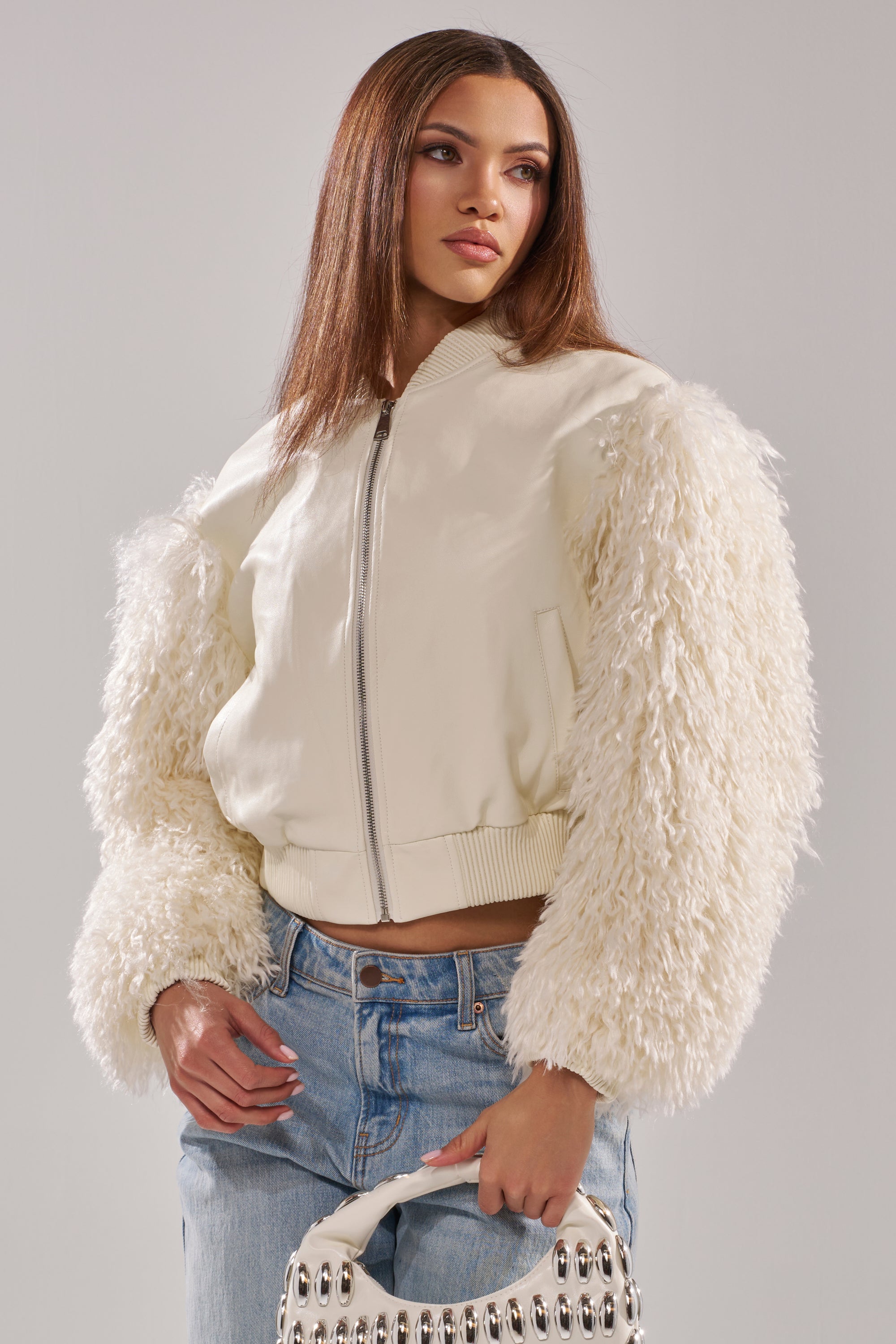 A woman wears the SO GOOD IT HURTS FUR SLEEVE BOMBER IN IVORY with fluffy sleeves and blue jeans, holding a white handbag with metallic accents. She has long, straight hair and stands against a plain light background. 3