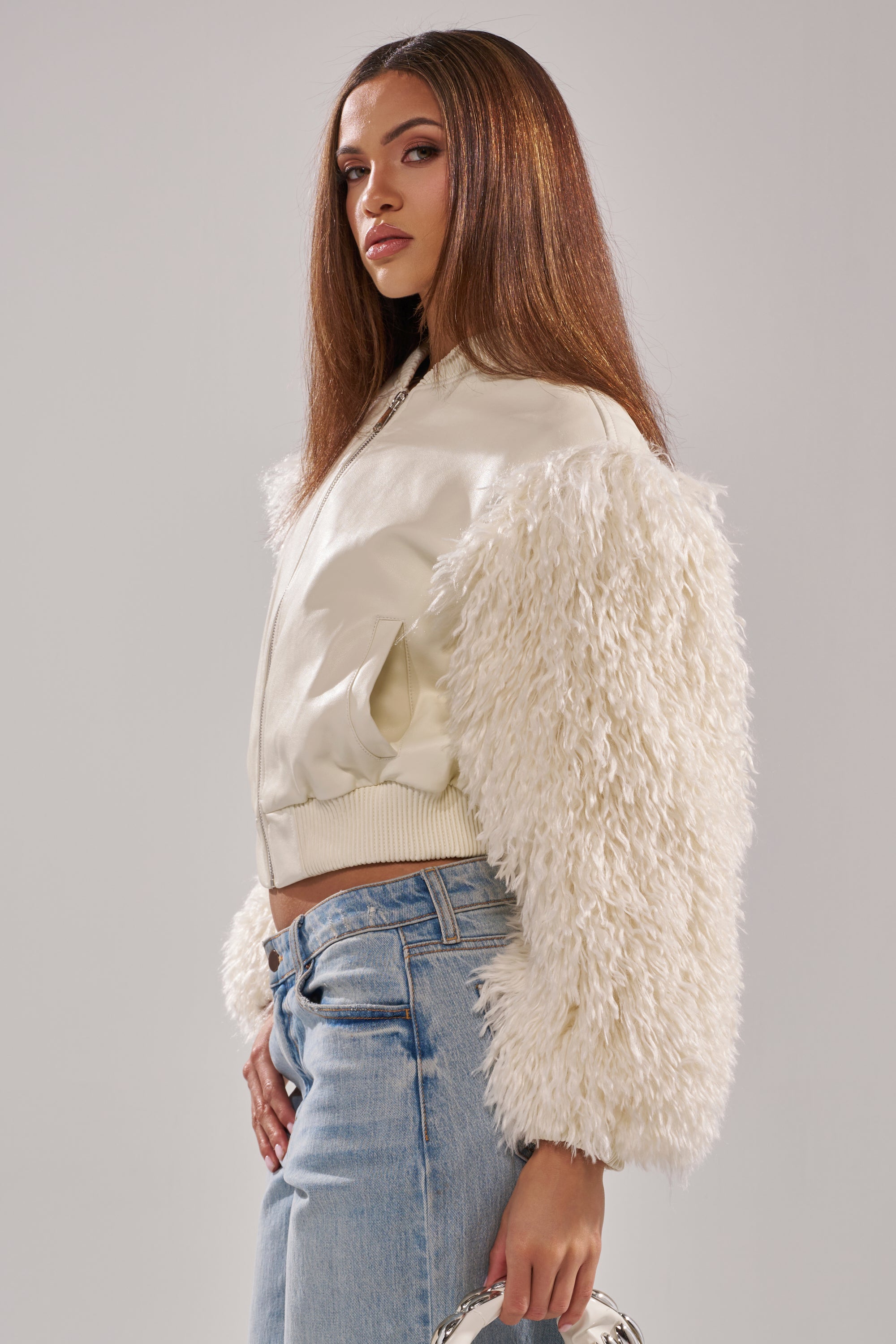 A woman with long straight hair stands sideways in light blue jeans, holding a small silver handbag and wearing the SO GOOD IT HURTS FUR SLEEVE BOMBER IN IVORY. 5
