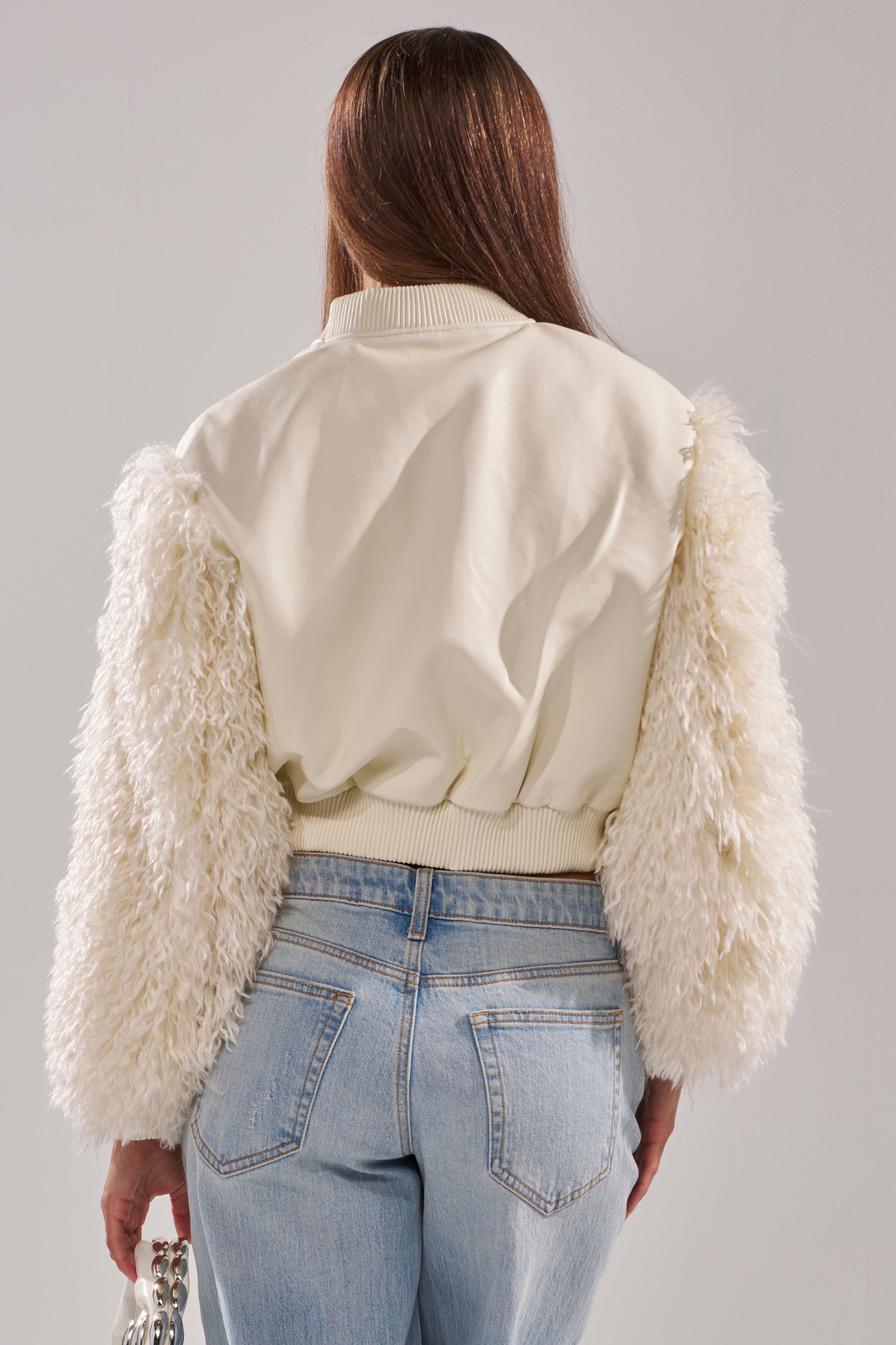 A person with long brown hair is shown from behind wearing the SO GOOD IT HURTS FUR SLEEVE BOMBER IN IVORY with textured, fluffy sleeves, paired with light blue jeans and holding a small silver handbag. 6