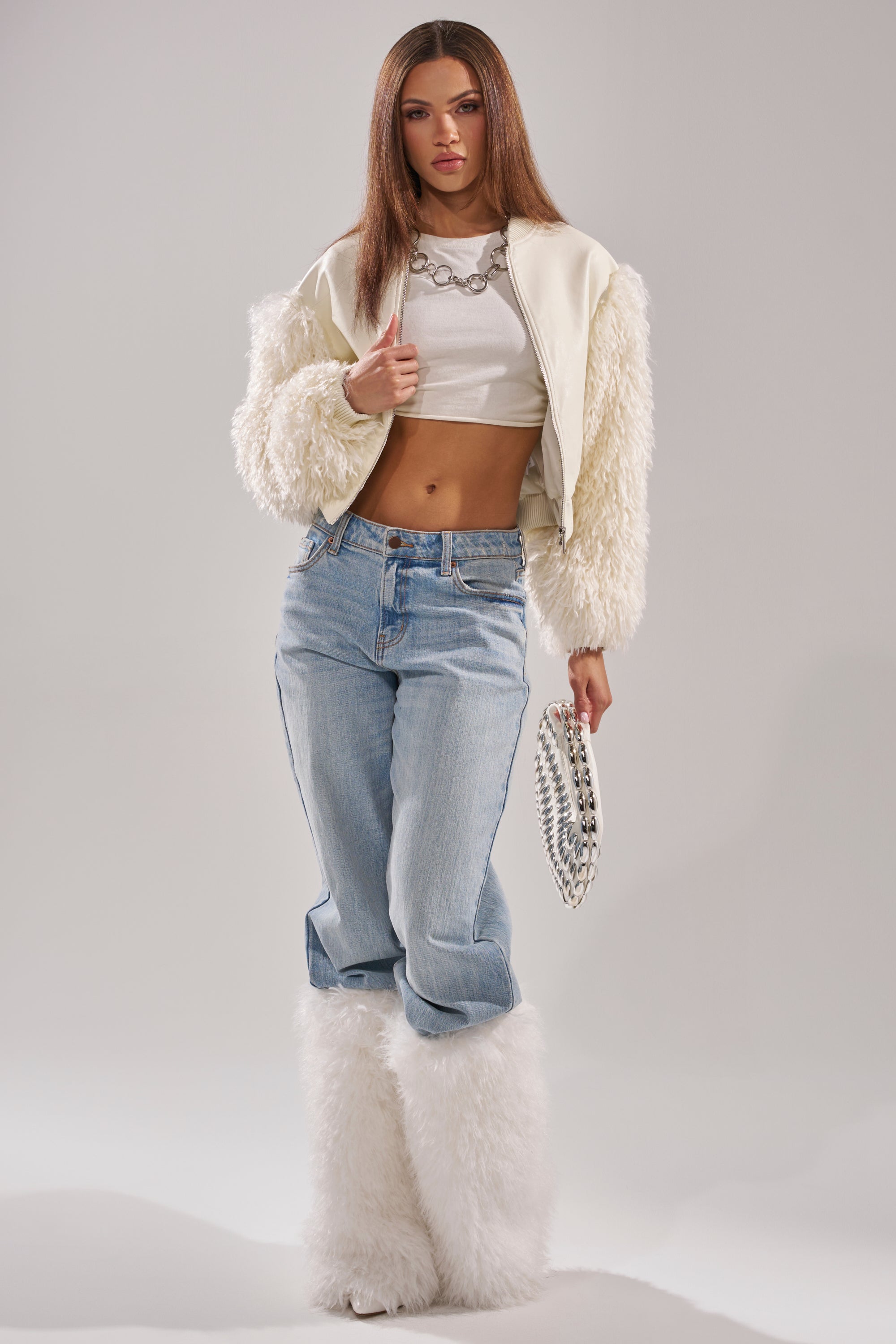A woman poses in the SO GOOD IT HURTS FUR SLEEVE BOMBER IN IVORY, a white crop top, light blue jeans, tall fluffy white boots, a chunky chain necklace, and holds a silver studded clutch against a plain light gray background. 8