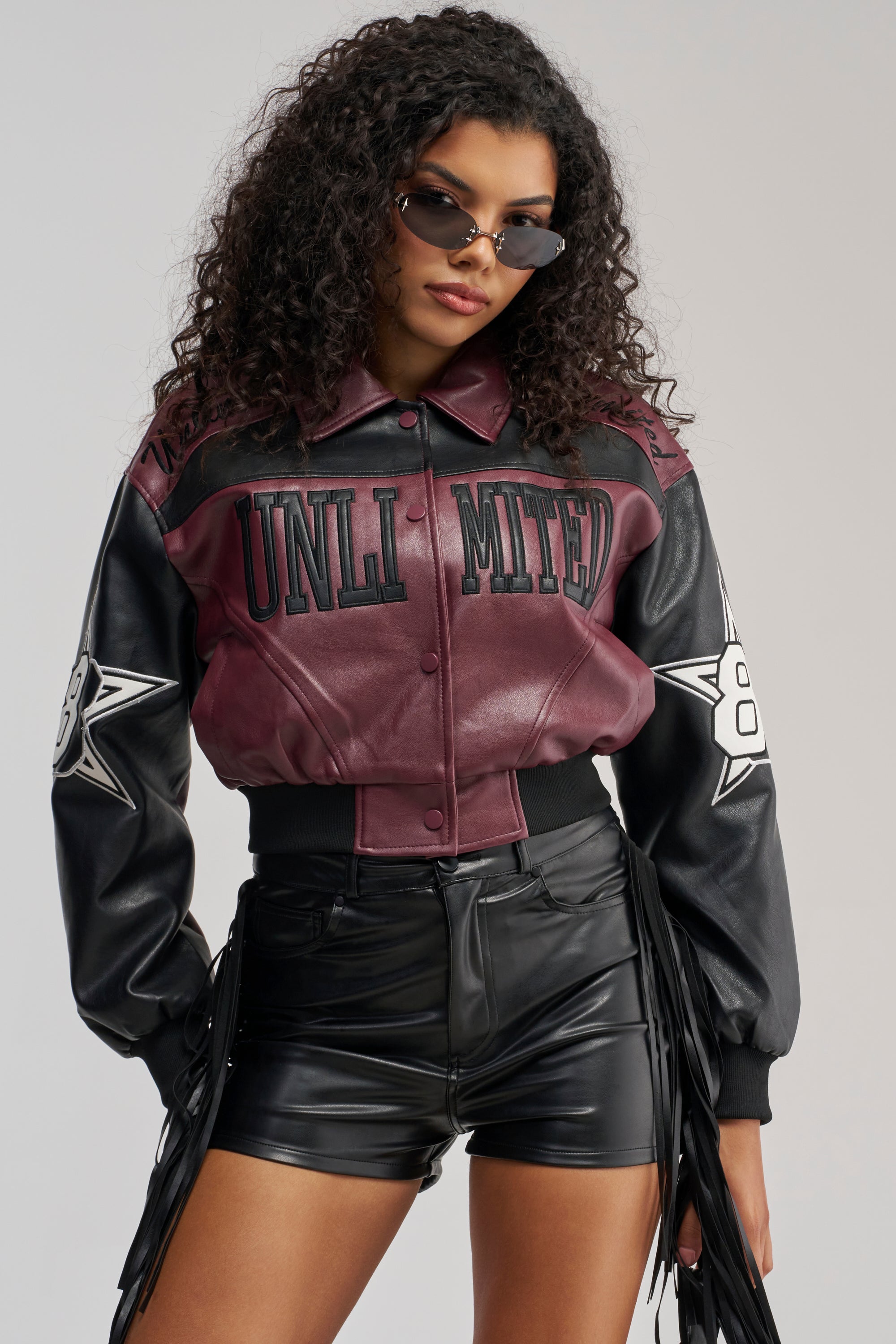 A woman in sunglasses poses confidently against a plain background, wearing the HARD TO FORGET MOTO BOMBER—a maroon and black vegan leather jacket featuring “UNLIMITED” text and star patches—paired with black fringe-detailed leather shorts.