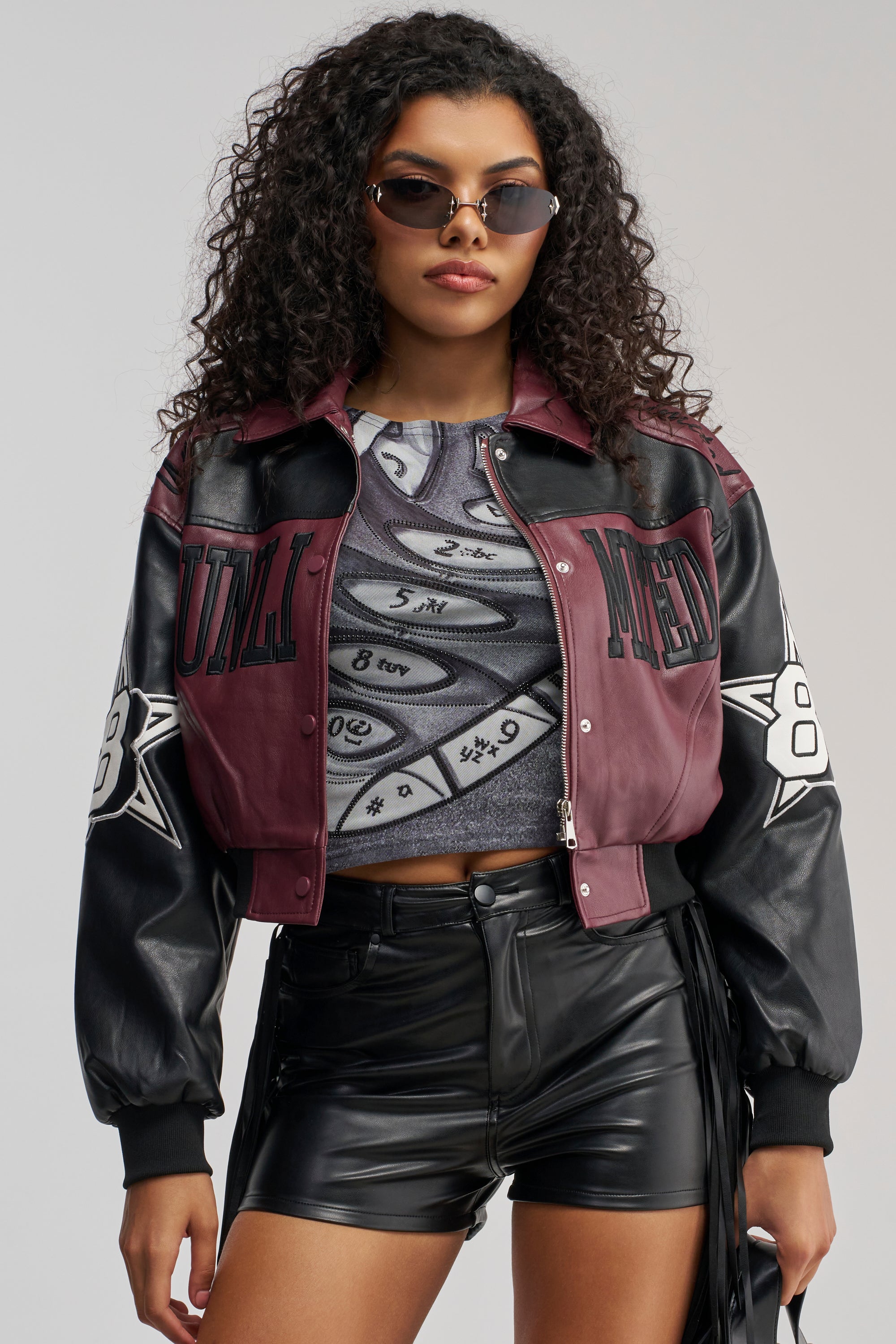 A woman with curly hair wears sunglasses and the HARD TO FORGET MOTO BOMBER—a varsity-inspired jacket in maroon and black—paired with a printed crop top and high-waisted black leather shorts, standing confidently against a plain background.