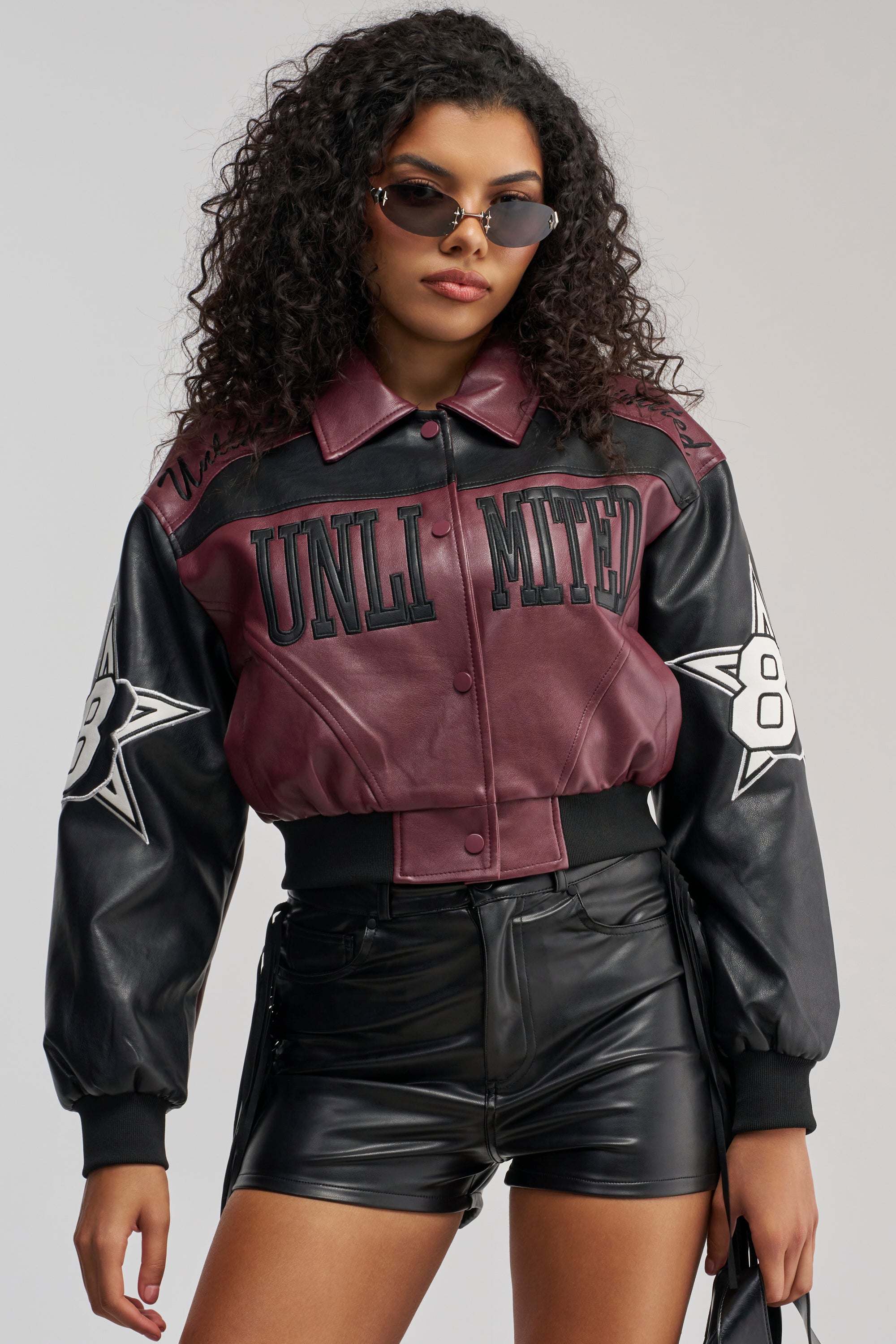 A woman with curly hair wears sunglasses and the HARD TO FORGET MOTO BOMBER—a maroon and black vegan leather varsity-inspired jacket with star and number patches—paired with black leather shorts, holding a matching bag against a plain background.