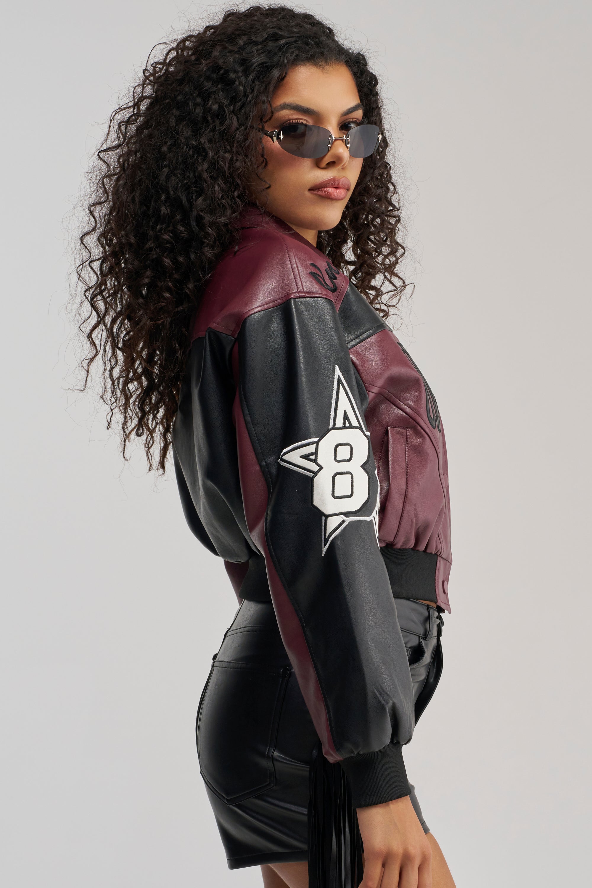 A woman with long curly hair wears sunglasses and the HARD TO FORGET MOTO BOMBER—a varsity-inspired, vegan leather jacket in black and burgundy featuring a white star and the number 8 on the sleeve—paired with black leather shorts, as she poses confidently against a neutral background.