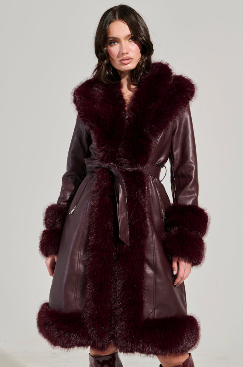 Against a plain background, a woman stands wearing the YUKON 2.0 FUR TRIM COAT, a luxurious dark burgundy faux leather coat featuring quilted lining and fur trim on the collar, cuffs, and hem, elegantly tied with a belt at the waist.