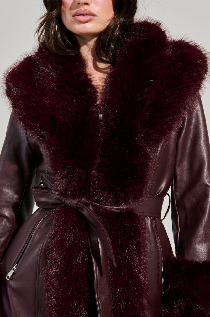 A woman stands against a neutral background, wearing the YUKON 2.0 FUR TRIM COAT in dark burgundy. The coat features a faux fur collar and quilted lining, with a tied belt that accentuates its luxurious and stylish appearance.