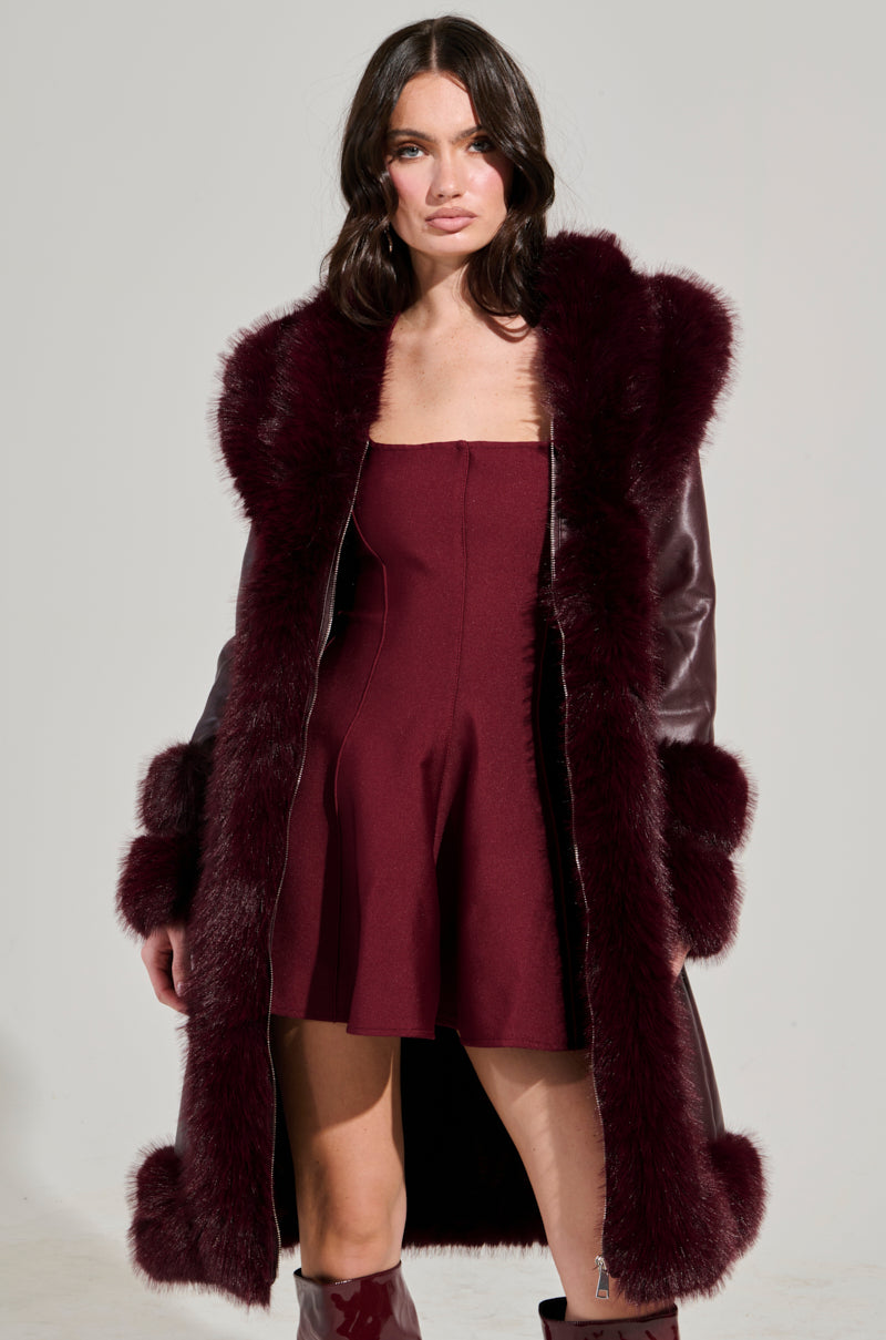 A person with long, dark hair poses confidently in a burgundy dress and the YUKON 2.0 FUR TRIM COAT, featuring a quilted lining, against a plain, light background.