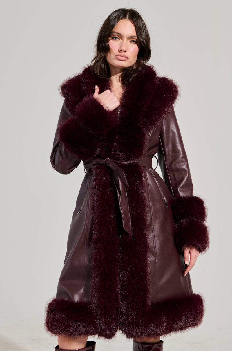 A woman showcases the YUKON 2.0 FUR TRIM COAT, a dark red, faux leather masterpiece with luxurious fur accents. This coat is adorned with a fur collar, cuffs, and hem, beautifully complemented by a quilted lining that adds elegance and depth. Standing against a simple gray backdrop, she gazes intently at the camera.