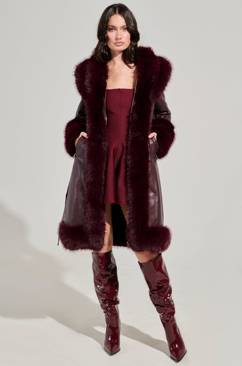 A woman stands confidently wearing the YUKON 2.0 Fur Trim Coat in burgundy, a matching strapless dress, and shiny knee-high boots. She poses against a plain background with her hands in her coat pockets.