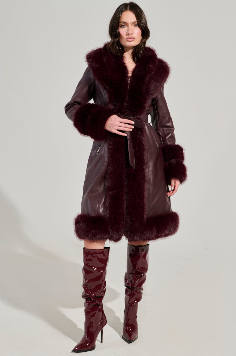 A person wearing the YUKON 2.0 FUR TRIM COAT, a dark purple piece with quilted lining, belt, and soft fur accents on the hems and cuffs, along with matching knee-high boots stands against a plain background.