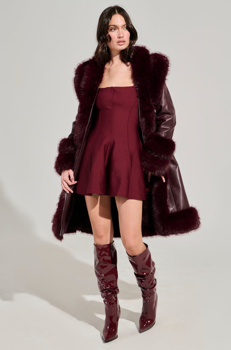 A woman is seen wearing the YUKON 2.0 FUR TRIM COAT, featuring a burgundy faux leather exterior with quilted lining and fur trim, over a strapless burgundy dress. Her long wavy hair complements her shiny burgundy knee-high boots against a plain light gray background.