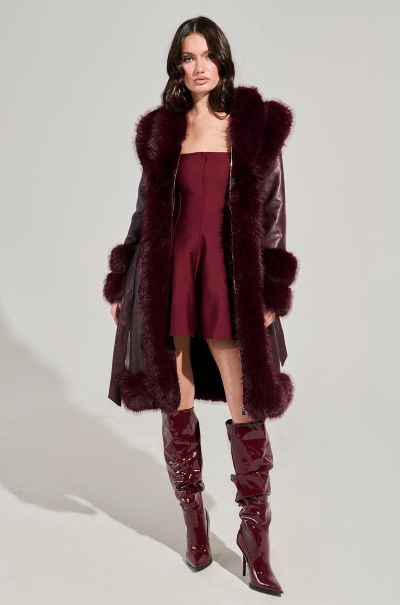 A woman stands wearing the YUKON 2.0 FUR TRIM COAT in burgundy faux leather, complete with quilted lining and fur trim, over a matching strapless dress and shiny knee-high boots. She has dark hair and a confident pose against a plain background.