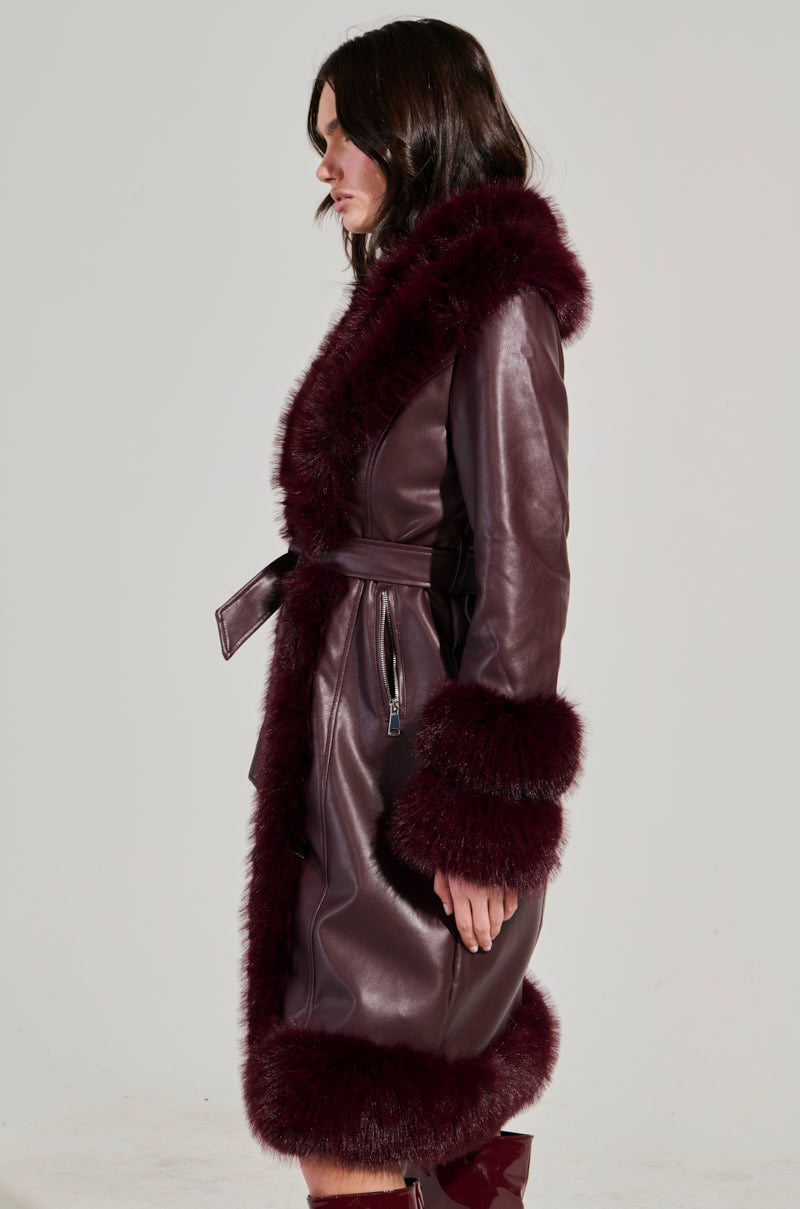 A person is wearing the YUKON 2.0 FUR TRIM COAT, a long, dark brown faux leather piece with deep burgundy fur trim adorning the hood, cuffs, and hem. With its quilted lining and waist tie, it beautifully complements their dark hair as they stand in profile against a plain background.