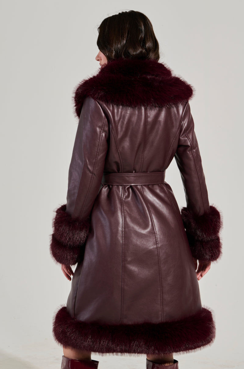 A person is wearing the YUKON 2.0 FUR TRIM COAT, a long faux leather coat in dark red with plush fur trim on the collar, cuffs, and hem. The coat is belted at the waist, and as they face away from the camera, it elegantly showcases the back of this stylish piece.