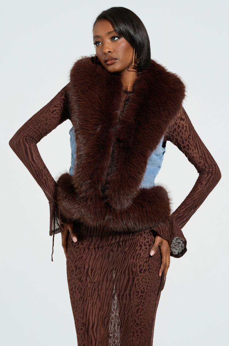 A woman stands confidently, exuding luxurious vibes in the ALICIA FUR TRIM DENIM VEST layered over a long-sleeved, animal-print dress. With one hand on her hip and the other relaxed, her smoothly styled hair completes the look against a plain light background.