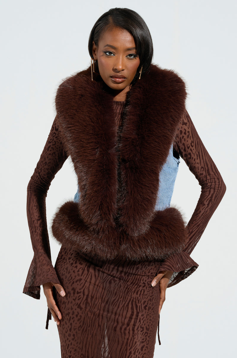 Against a light grey backdrop, a model radiates luxury wearing the ALICIA FUR TRIM DENIM VEST over a chic brown animal print dress. The ensemble is completed with flared sleeves for added style and long earrings that highlight her confident expression.