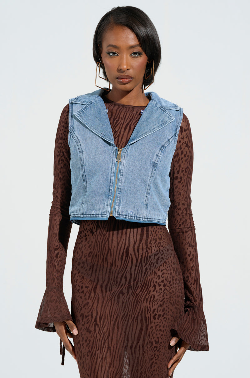 A person dressed in the ALICIA FUR TRIM DENIM VEST over a long-sleeved, brown patterned dress. Their shoulder-length hair pairs beautifully with large hoop earrings, creating a luxurious look against the plain white background.
