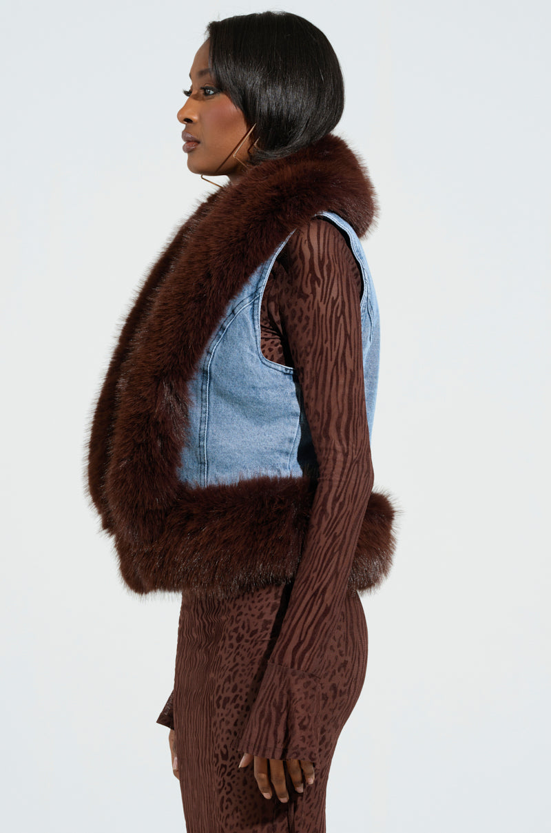 A person with long dark hair is wearing a brown patterned dress and the ALICIA FUR TRIM DENIM VEST. They stand in profile against a plain background, exuding subtle luxury vibes.