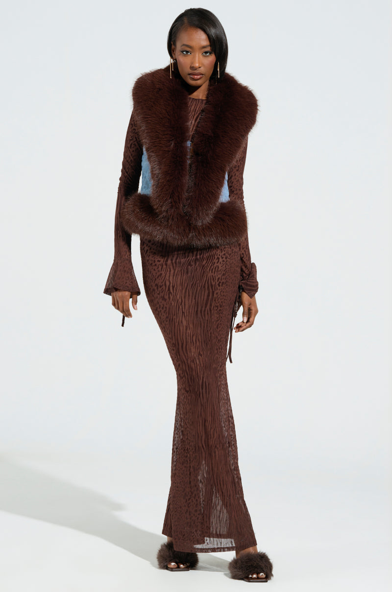 A woman is wearing a long, textured brown dress paired with the ALICIA FUR TRIM DENIM VEST, featuring sherpa lining and slippers. She stands against a plain white background, her hair down and expression neutral, exuding an aura of subtle luxury.
