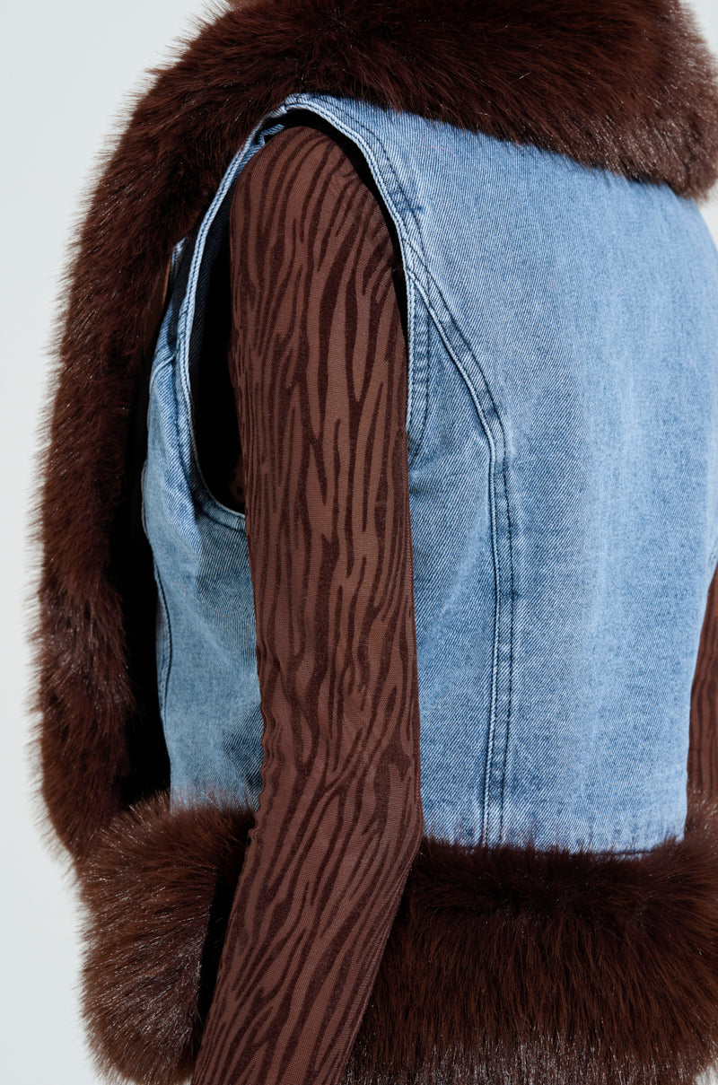 A model showcases a brown striped long-sleeve top layered with the ALICIA FUR TRIM DENIM VEST, exuding luxury vibes. The vest is accentuated by brown fur trim on the shoulders and hem, with the focus on the upper body drawing attention to the contrasting textures of the ensemble.
