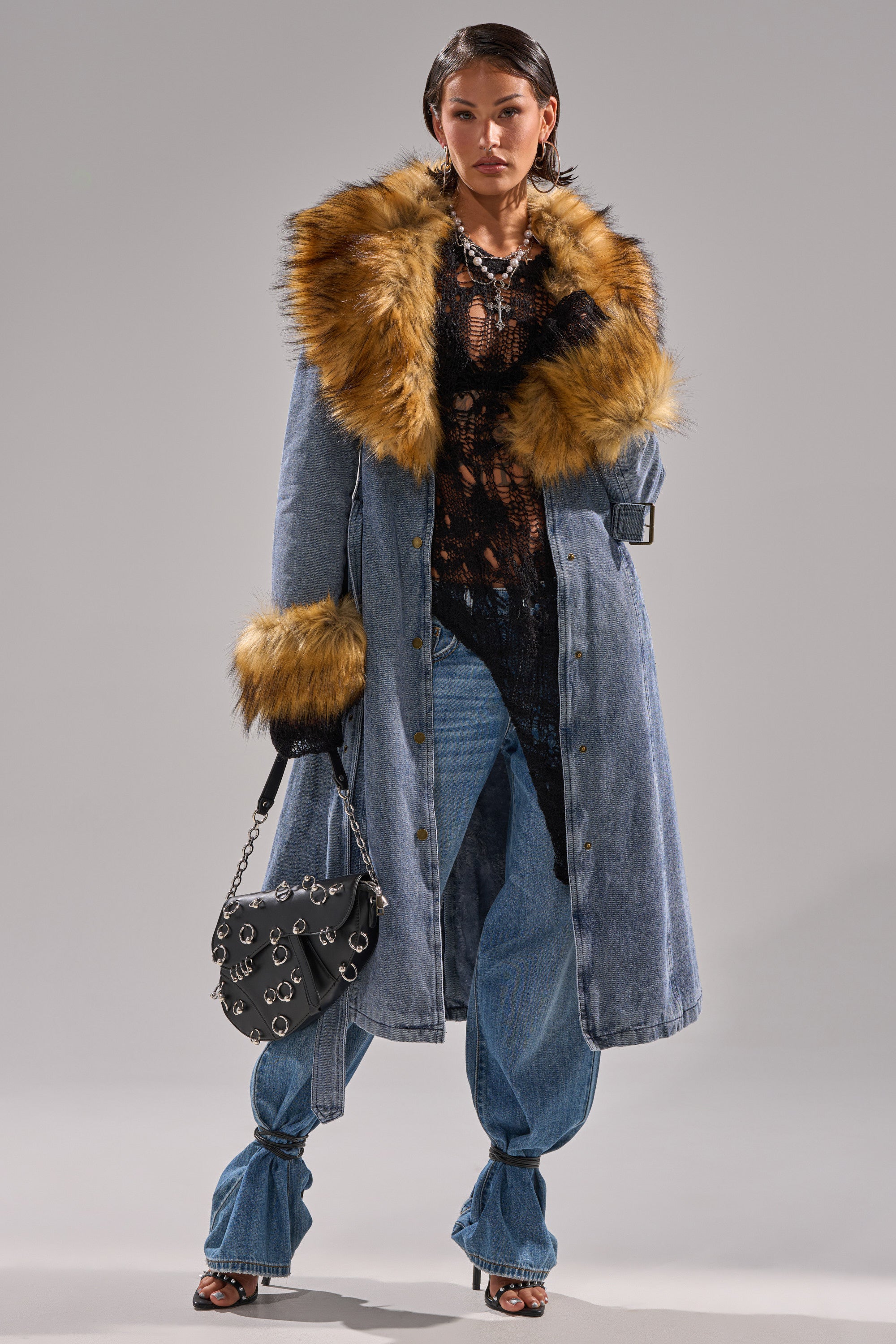 A woman models the KAYA FUR TRIM DENIM TRENCH with a large faux fur collar and cuffs, styled over a sheer black top and blue jeans. She accessorizes with a silver-ringed black handbag, heeled sandals, and layered necklaces.
