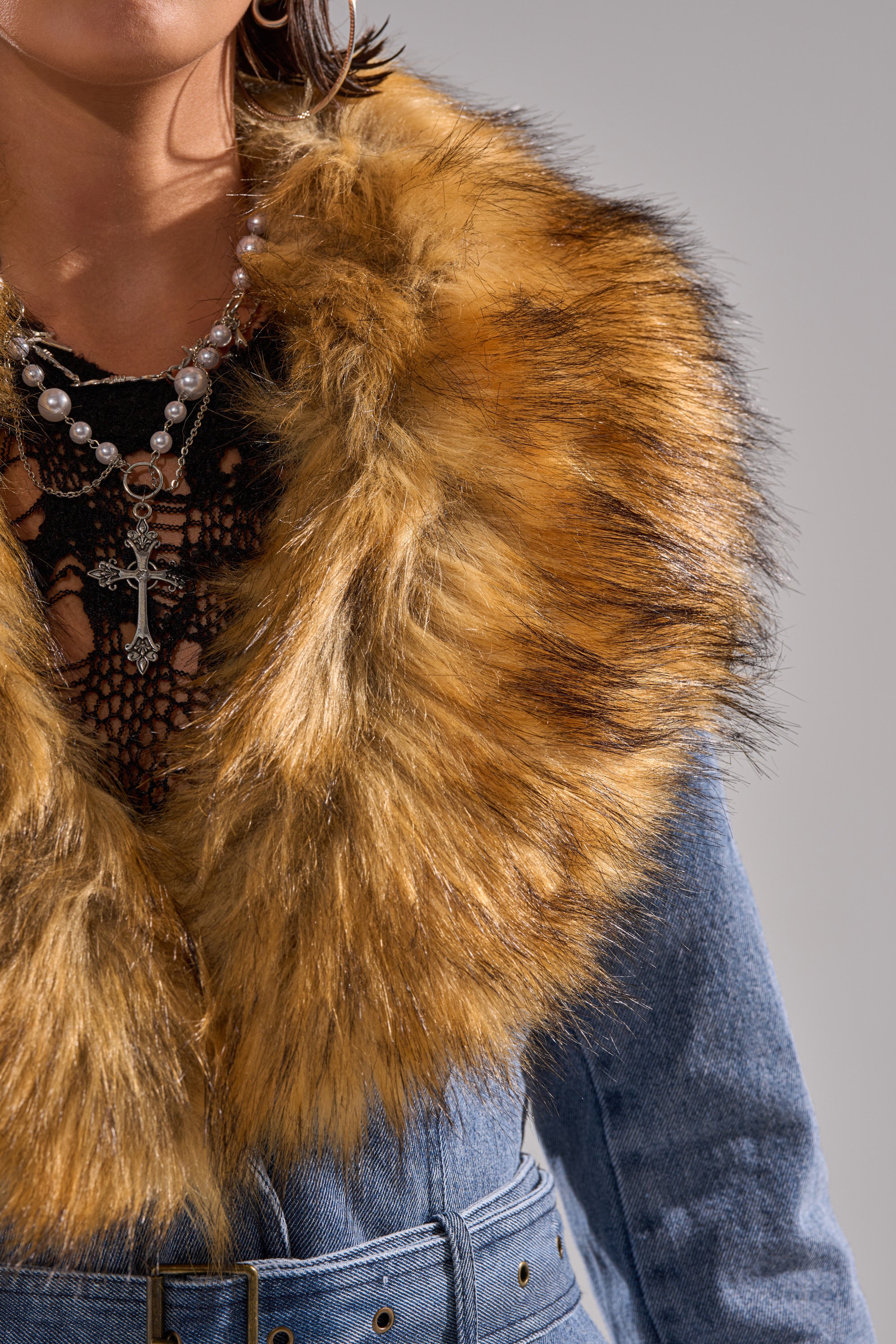 A person shown from shoulders to waist wears the KAYA FUR TRIM DENIM TRENCH, featuring blue denim and a large faux fur collar, styled with layered necklaces (one with a cross) and a black crocheted top underneath.