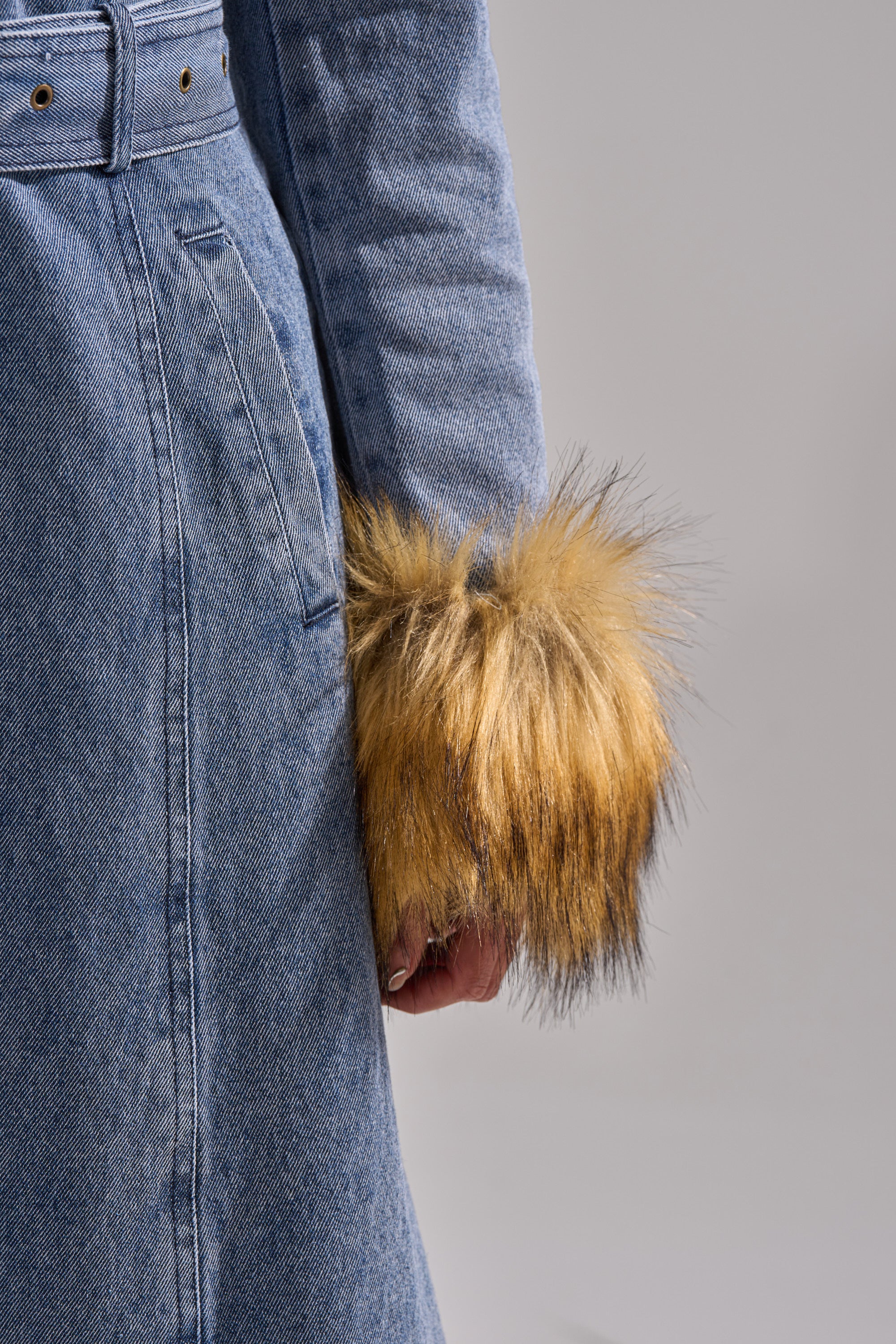 A person wears the KAYA FUR TRIM DENIM TRENCH, a blue denim coat with a belt and fluffy brown and black faux fur cuffs. One arm is visible, showing the sleeve detail against a plain light background.