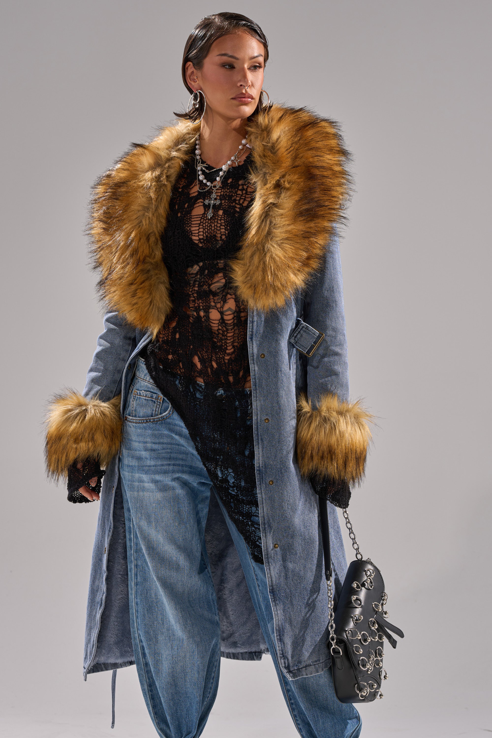 A woman models the KAYA FUR TRIM DENIM TRENCH, featuring a light blue wash with brown faux fur collar and cuffs, styled with a black lace top, loose jeans, and a studded black handbag with silver chain strap.