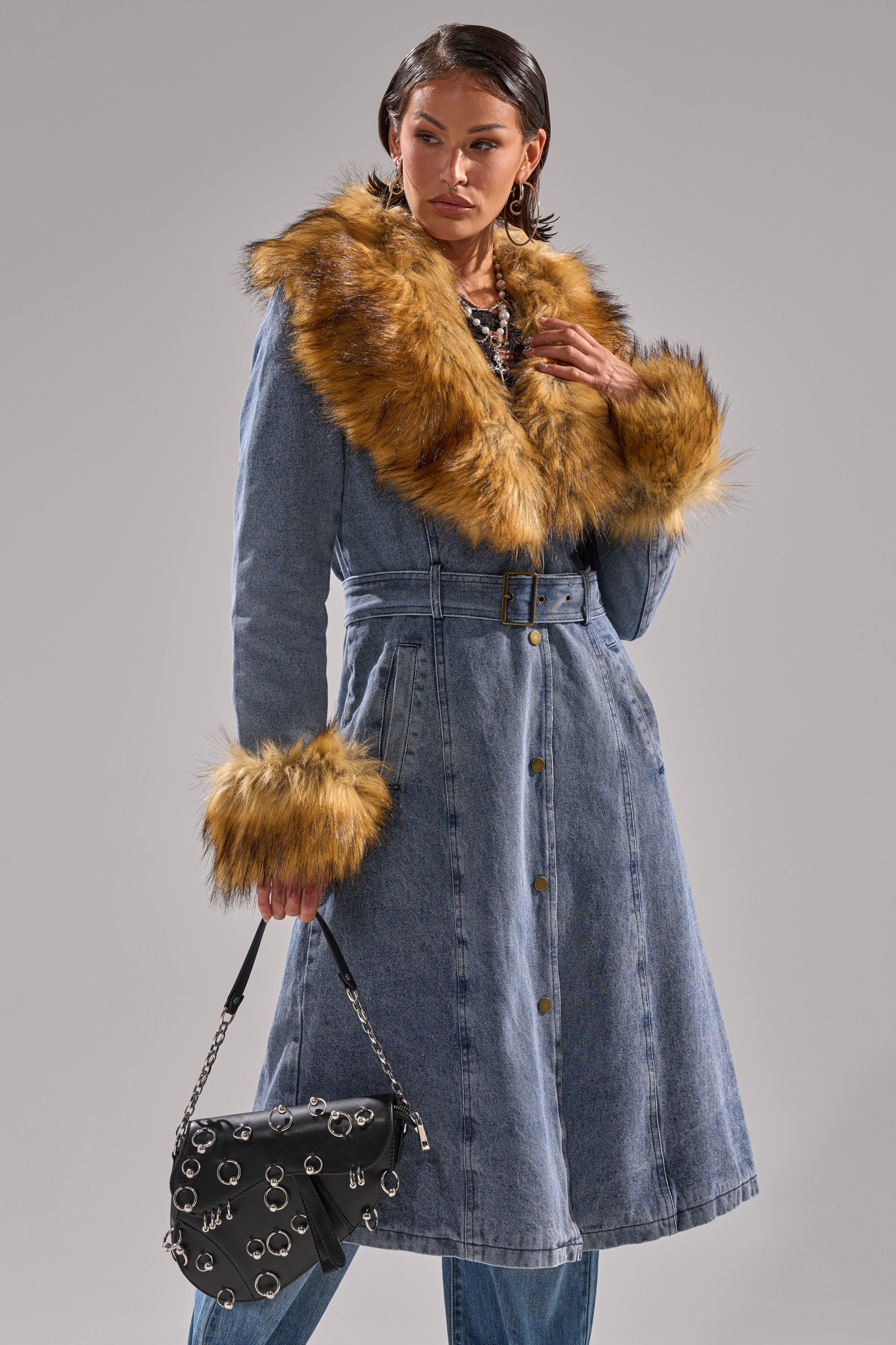A woman wears the KAYA FUR TRIM DENIM TRENCH with a faux fur collar and large brown cuffs, paired with blue jeans and holding a black handbag with metal rings, standing against a plain gray background.