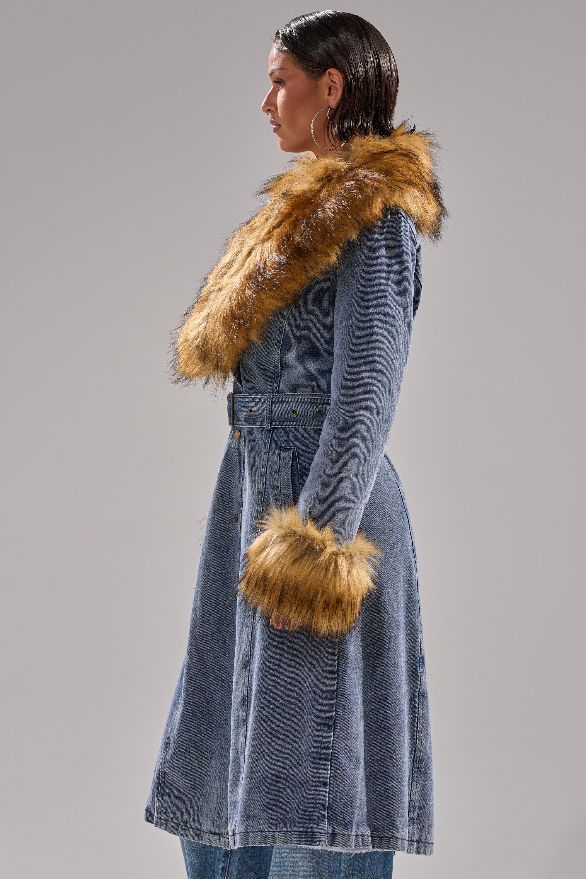 A person in profile models the KAYA FUR TRIM DENIM TRENCH, featuring a brown faux fur collar and cuffs, set against a plain gray background.
