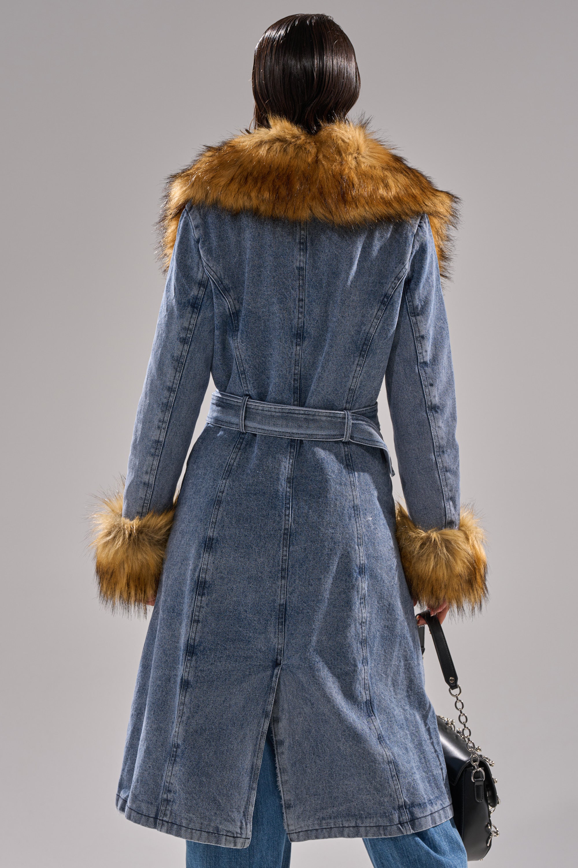 A person, back to the camera, wears the KAYA FUR TRIM DENIM TRENCH—a long, belted denim coat with faux fur collar and cuffs—while holding a small black handbag.