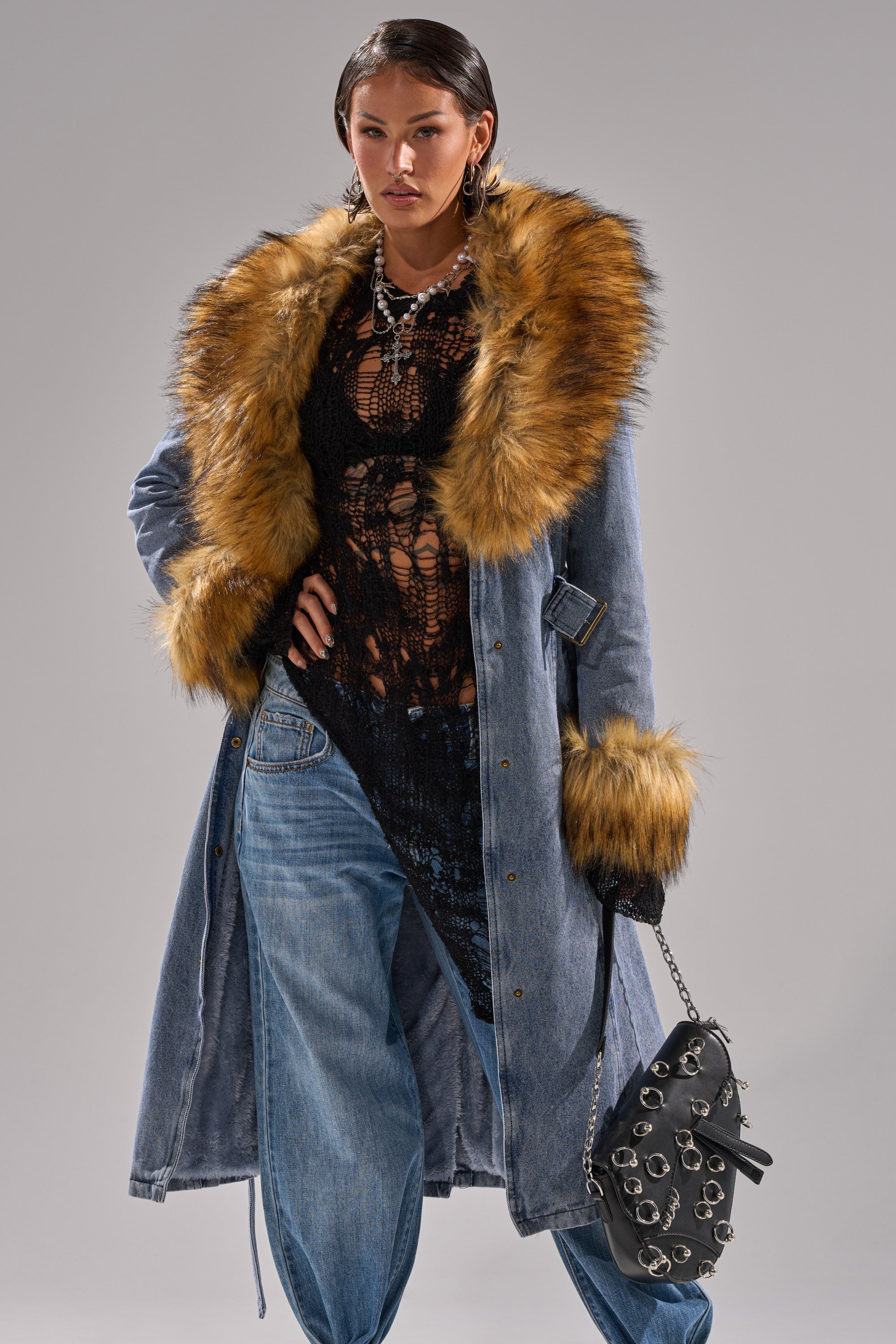 A woman confidently models the KAYA FUR TRIM DENIM TRENCH—a long blue denim coat with a bold faux fur collar and cuffs—paired with a black lace top, loose blue jeans, and a black handbag with silver ring accents.