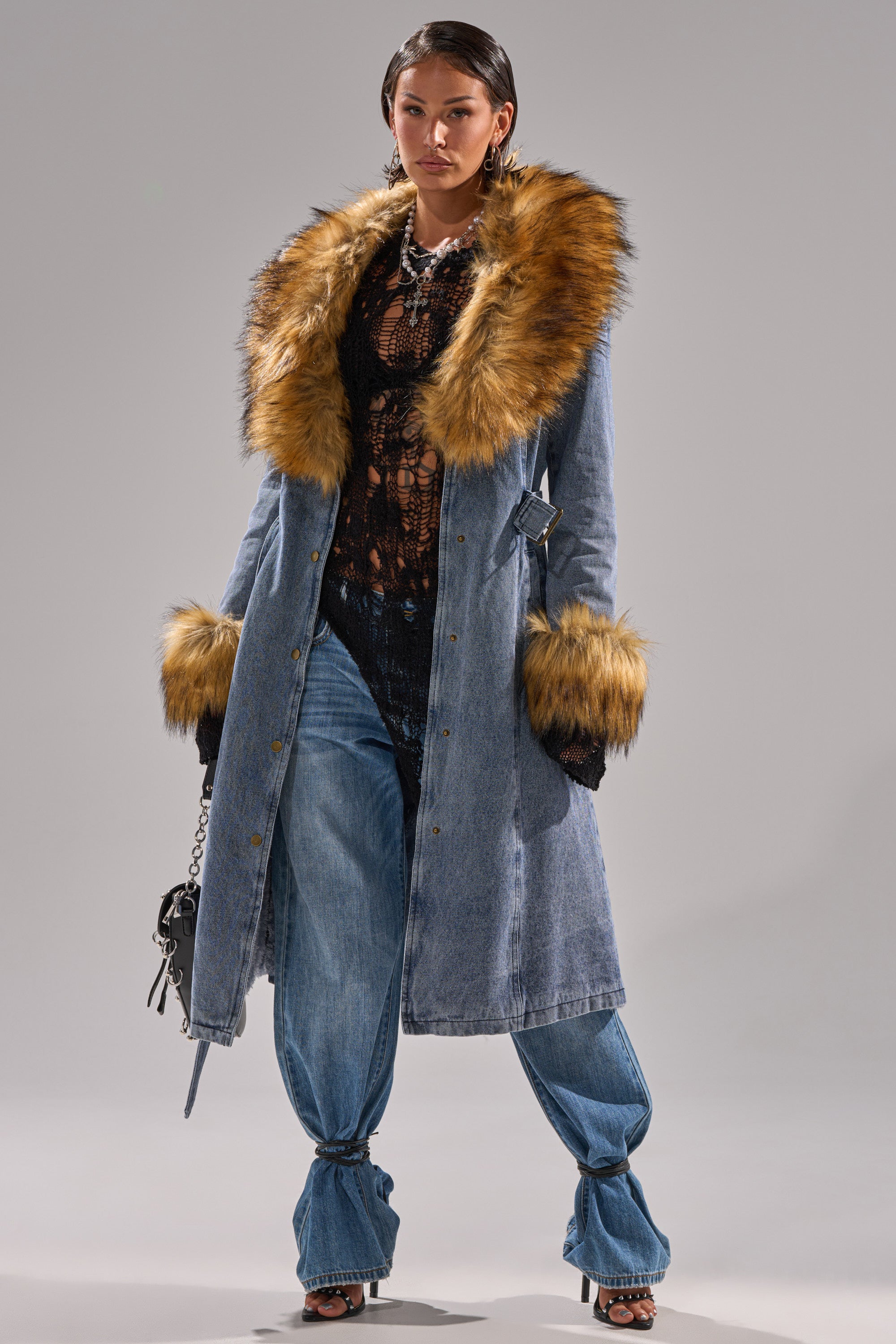A woman wears the KAYA FUR TRIM DENIM TRENCH, featuring a faux fur collar and cuffs, over a black lace top and wide-leg jeans. She accessorizes with a small bag and high-heeled shoes.