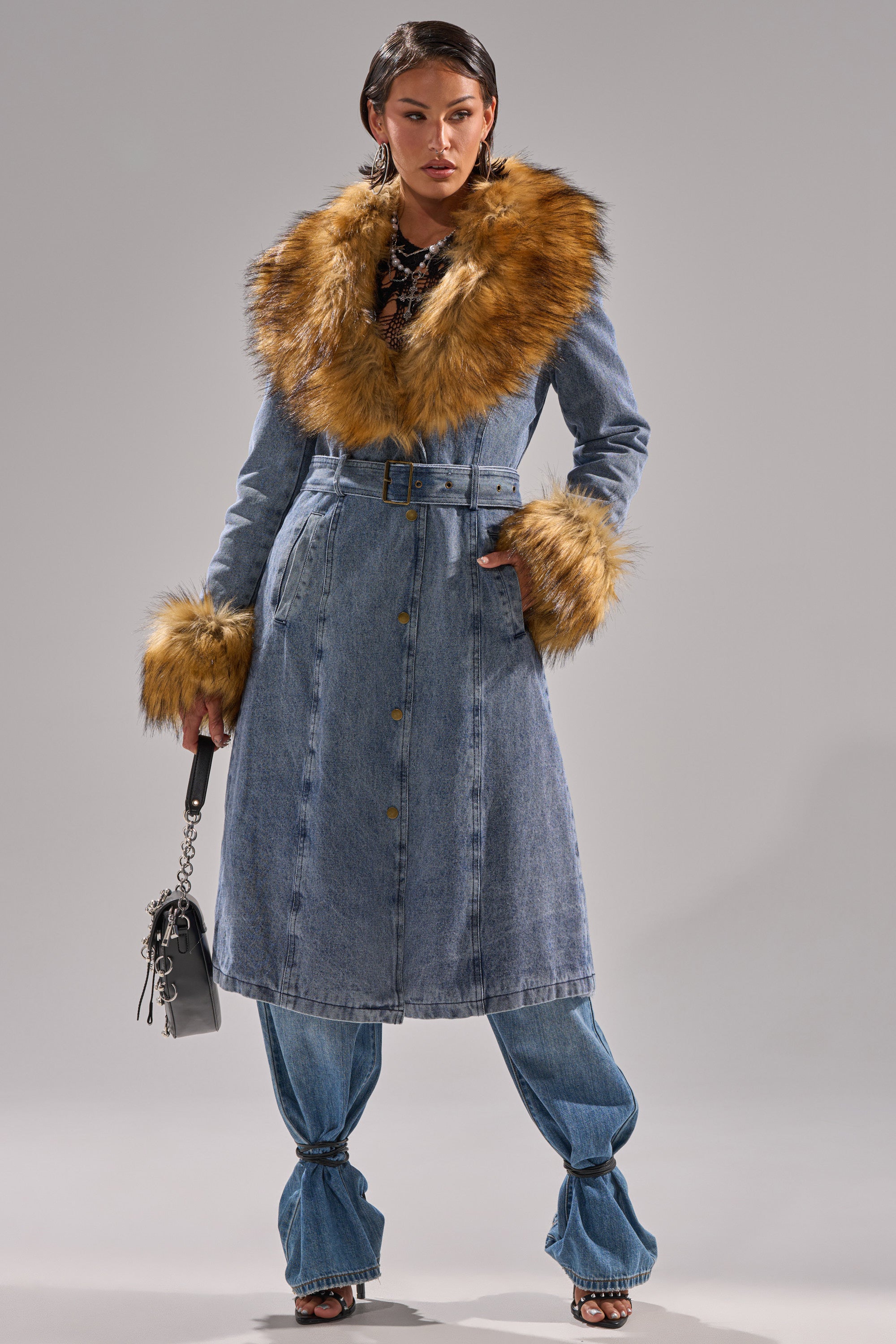 Wearing the KAYA FUR TRIM DENIM TRENCH with a large brown faux fur collar and cuffs, blue jeans, black heels, and holding a small black handbag, a woman stands confidently against a plain gray background.