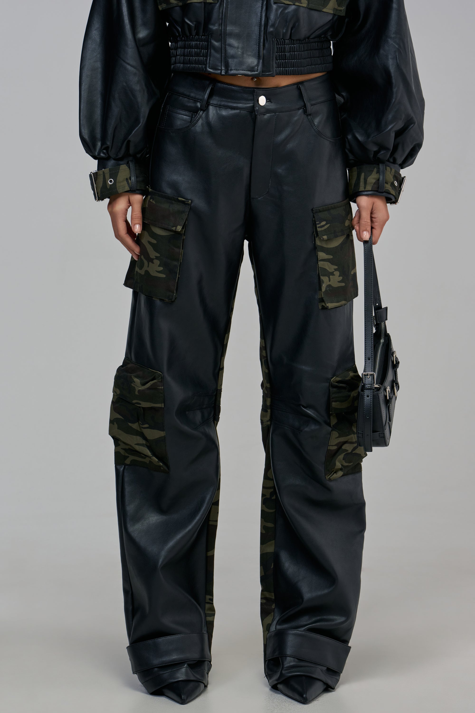 DOWN AND DIRTY FAUX LEATHER CAMO CARGO TROUSER in black faux leather with camouflage pattern panels on the pockets and legs, worn with a matching jacket and carrying a small black handbag.