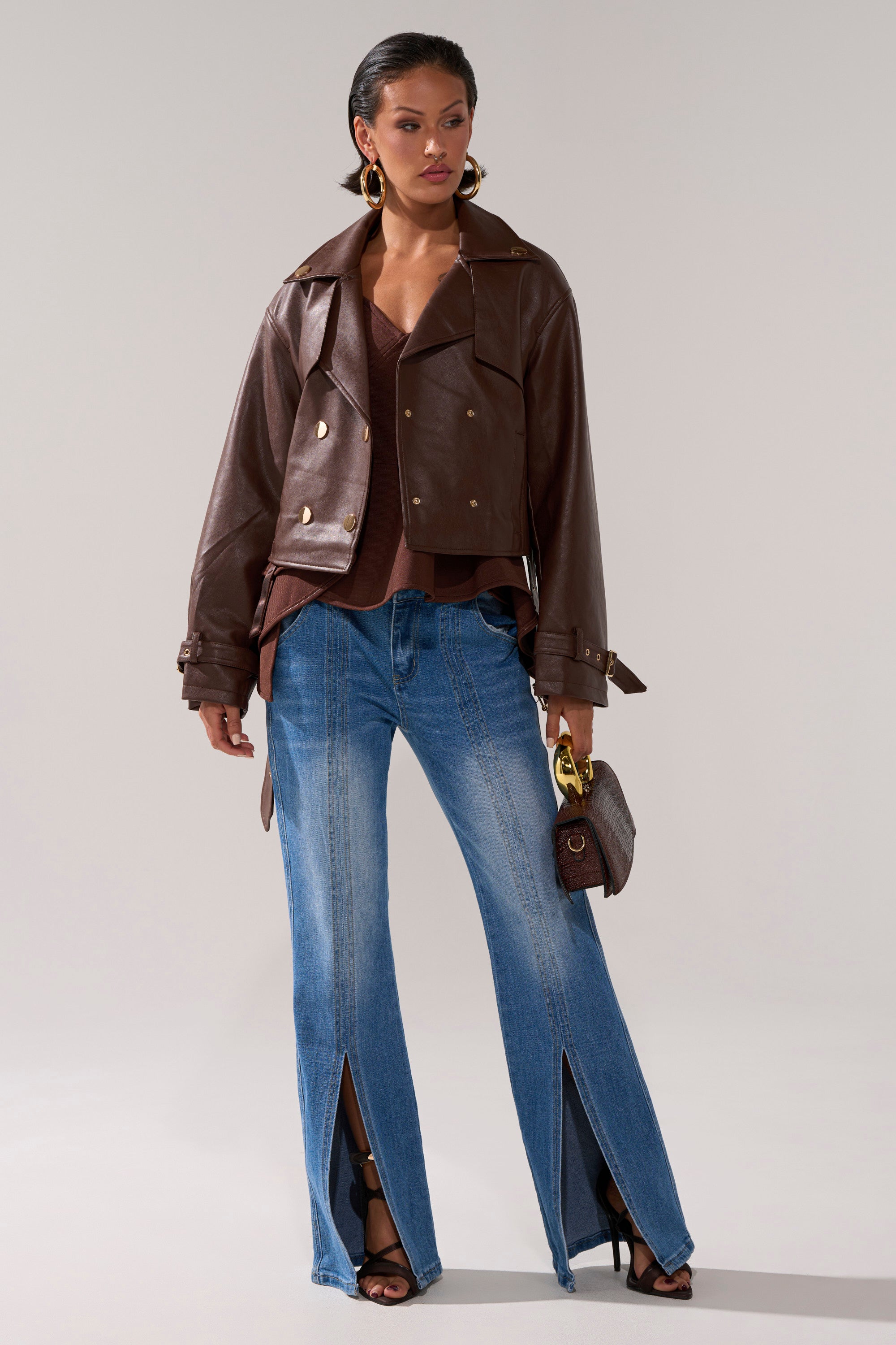 A woman wears the AKIRA Label LINA TRENCH MOTO faux leather cropped jacket, a brown top, blue jeans with front ankle slits, black heeled sandals, large hoop earrings, and carries a small brown handbag.