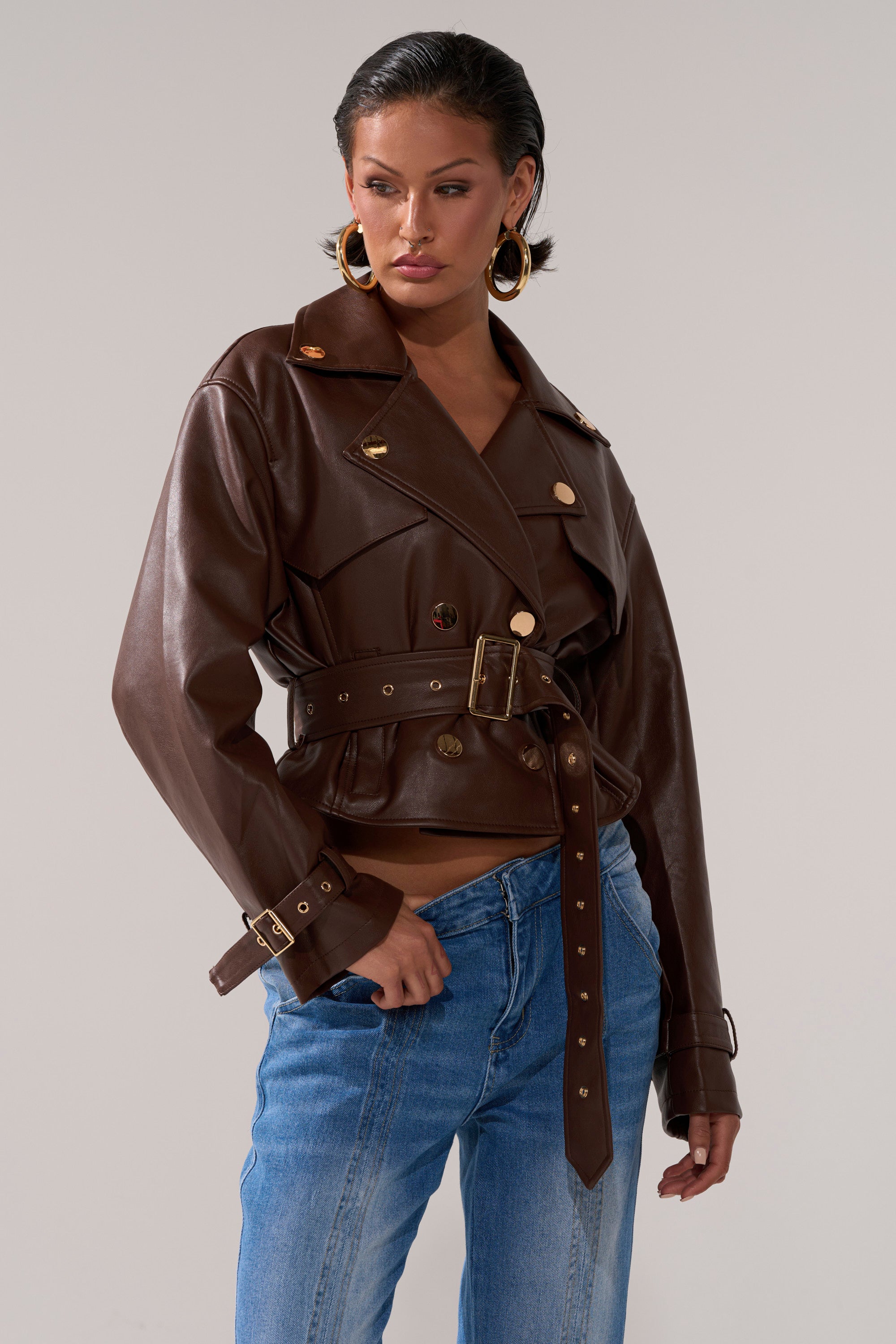 A woman wears the LINA TRENCH MOTO, a brown moto trench coat with gold buttons, styled with blue jeans. She poses with one hand in her pocket, sporting slicked-back hair and large gold hoop earrings against a plain background.