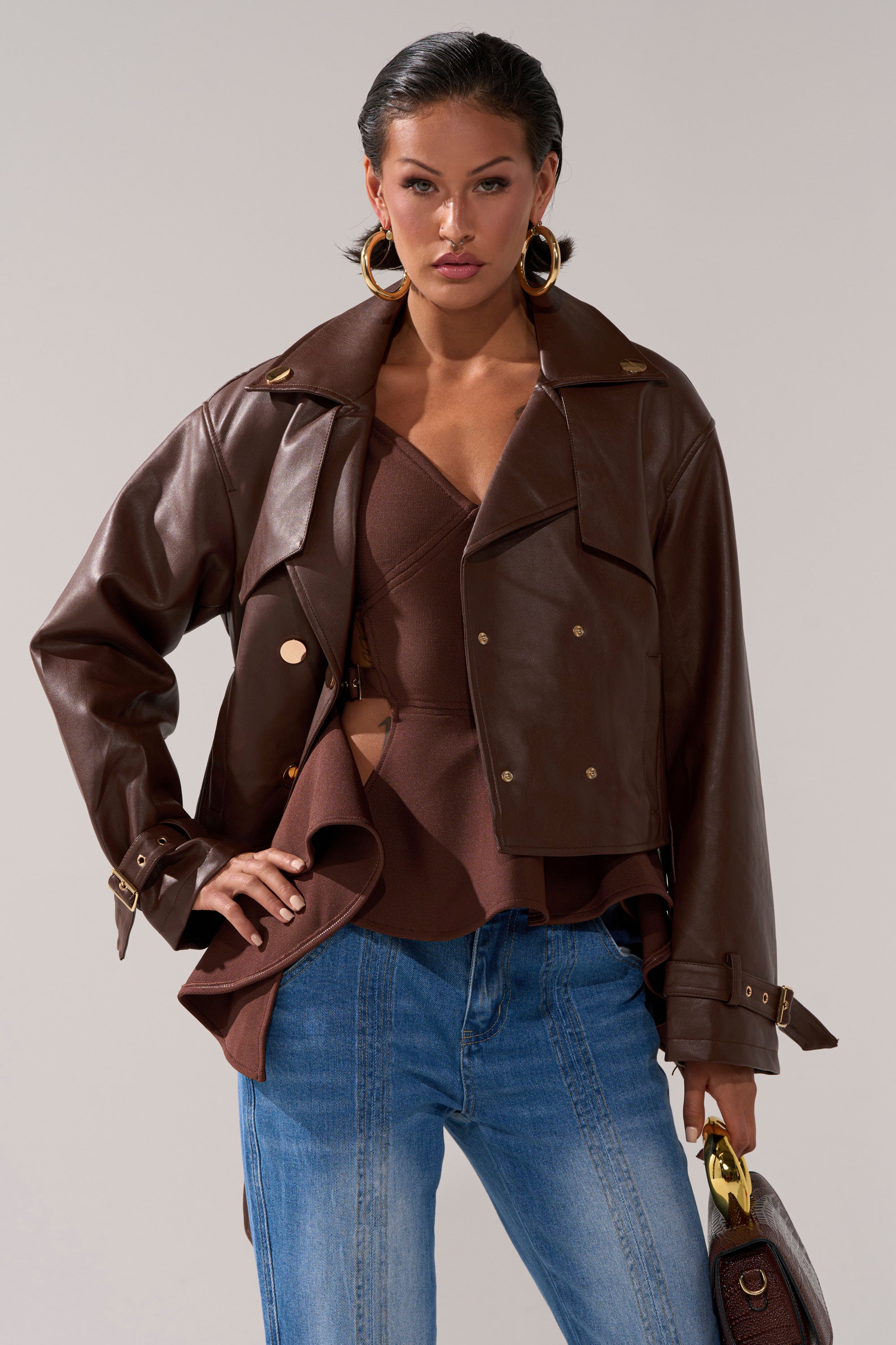 A woman poses confidently in the AKIRA Label LINA TRENCH MOTO faux leather cropped jacket over a brown ruffled top, blue jeans, large gold hoop earrings, and holding a brown handbag against a neutral background.