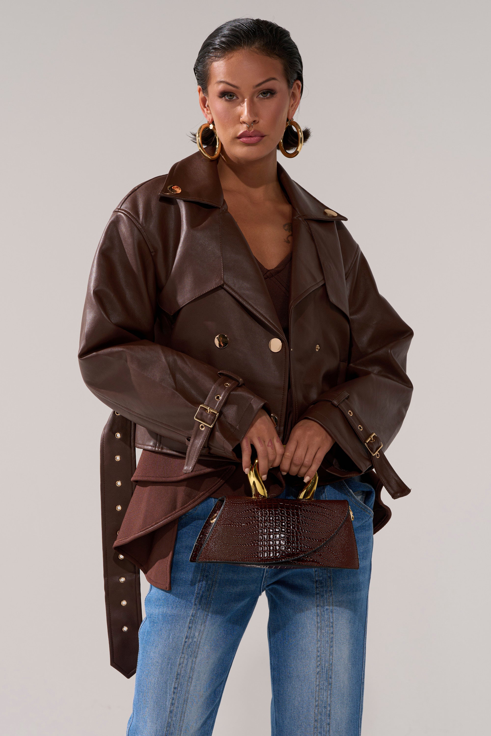Wearing the LINA TRENCH MOTO belted cropped jacket, hoop earrings, and blue jeans, a woman stands holding a textured brown handbag with gold handles against a light gray background.
