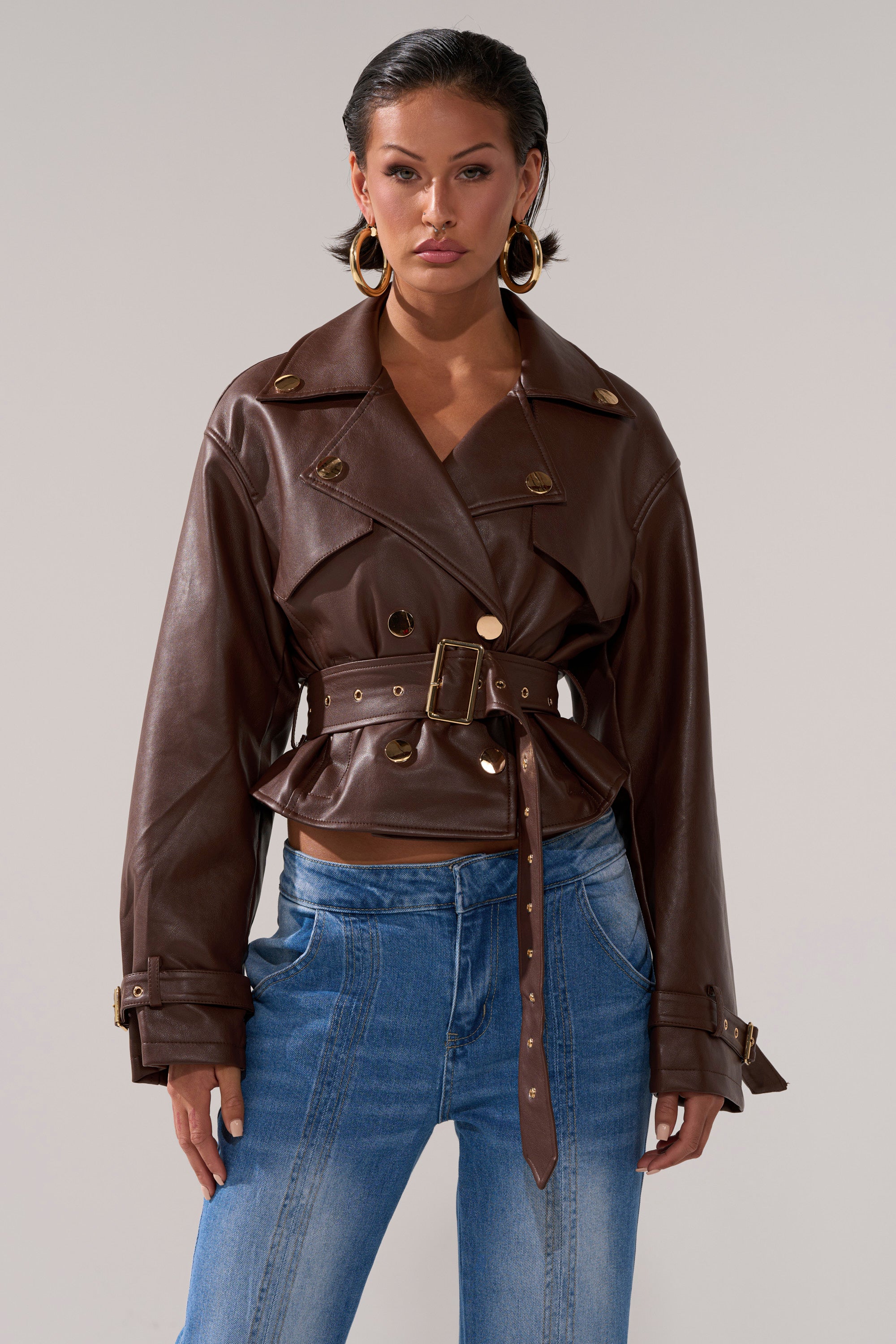 A woman stands facing forward against a plain background, wearing the LINA TRENCH MOTO—a belted cropped jacket with gold buttons—paired with blue high-waisted jeans and gold hoop earrings, her expression serious.