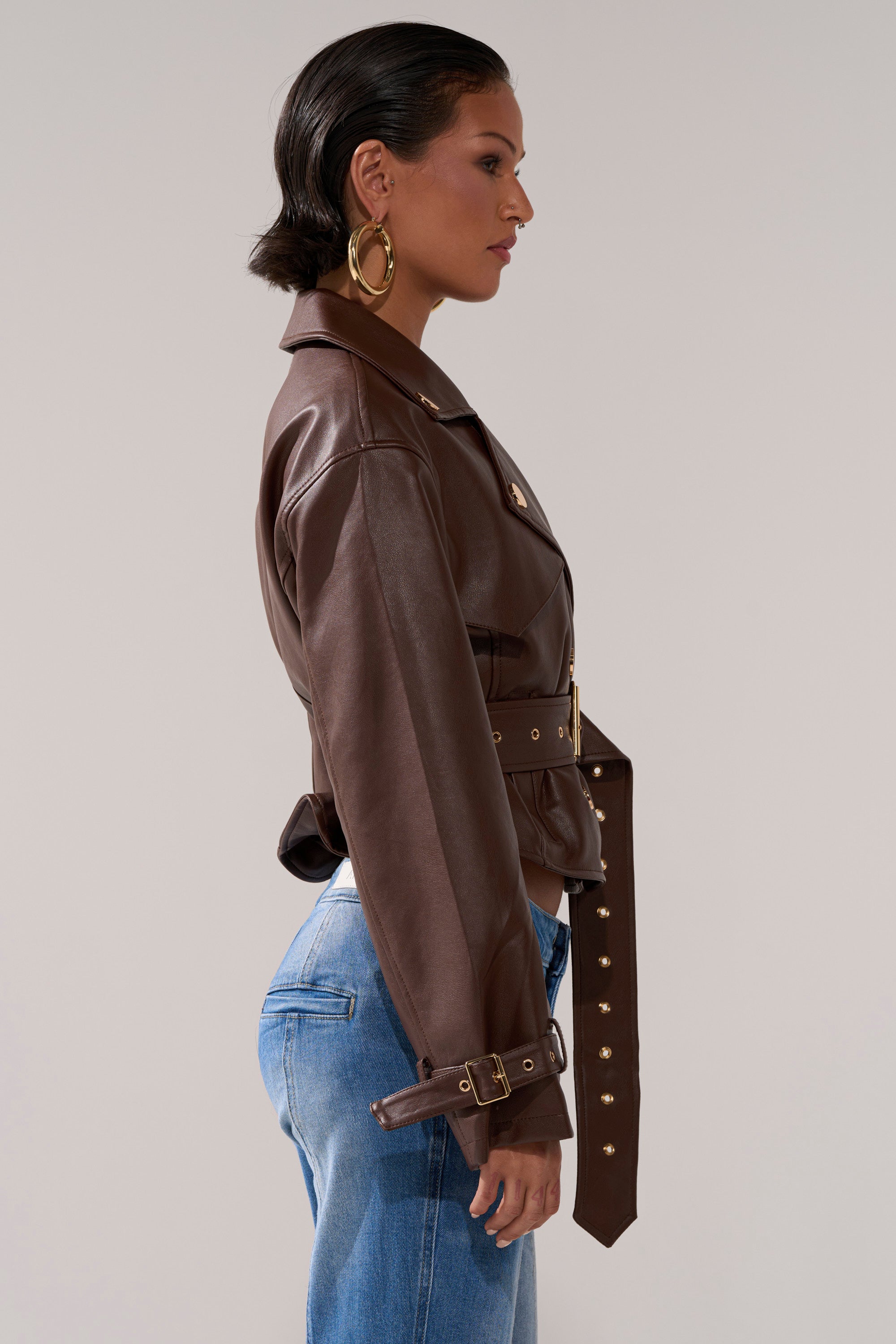 A woman in profile wears the LINA TRENCH MOTO, styled with large gold hoop earrings and blue jeans, standing against a plain light background.