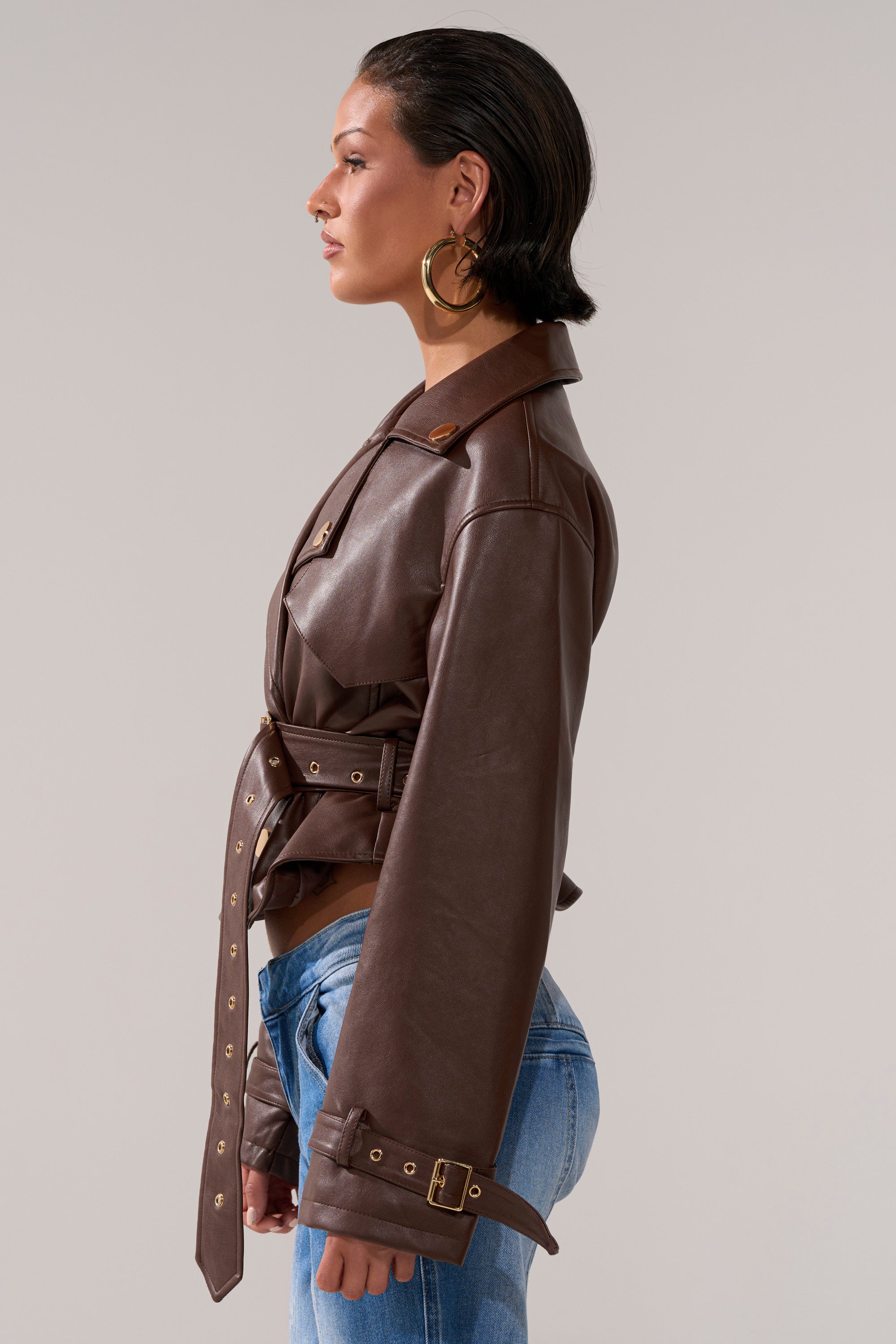 A person with short dark hair, large gold hoop earrings, and blue jeans wears the LINA TRENCH MOTO—a brown belted cropped jacket with buckled cuffs—standing in profile against a neutral background.