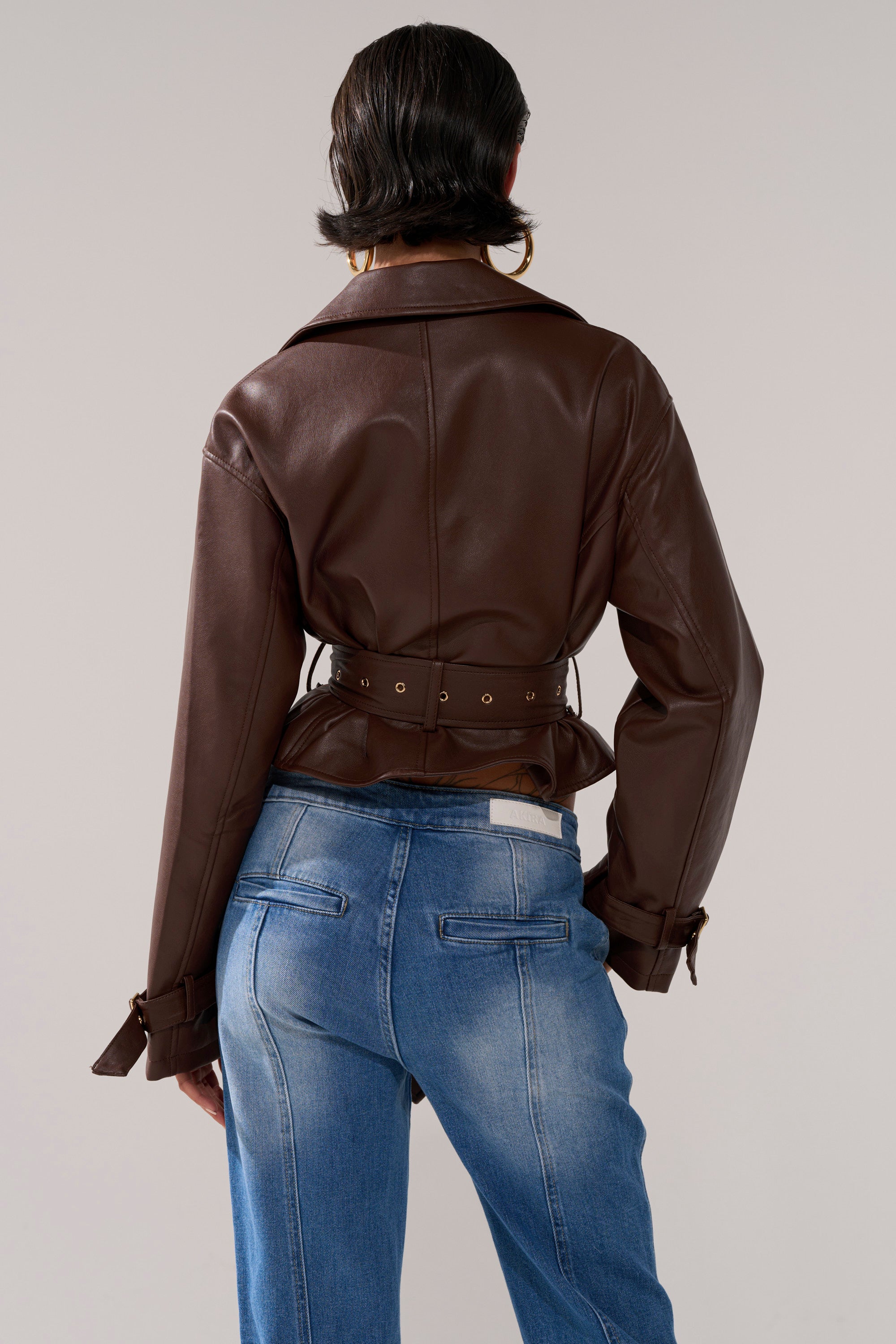 A person with short dark hair, wearing the LINA TRENCH MOTO jacket and blue jeans, stands facing away from the camera against a plain background.