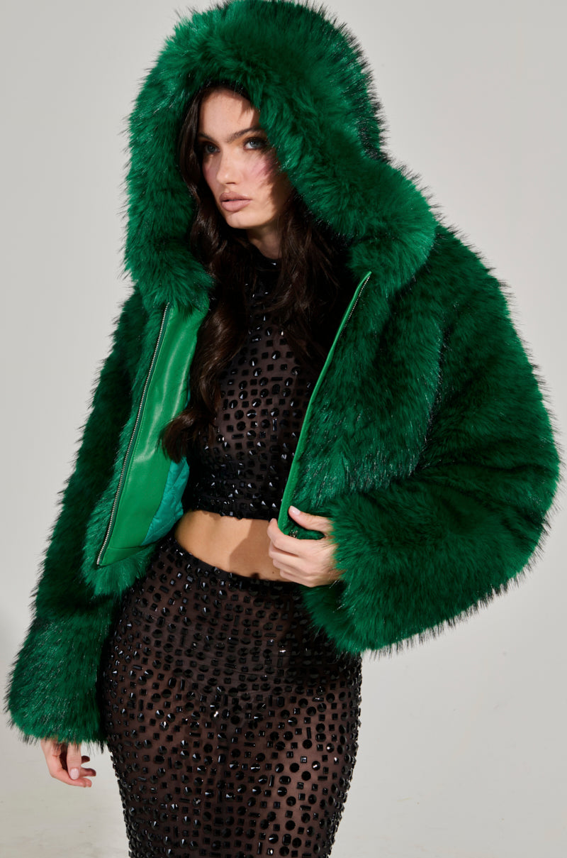 A person models a bold fashion outfit: the LUCKY HOODED FUR BOMBER in green, featuring the hood up, paired with a fitted black dress embellished with shiny details. The neutral background accentuates the vibrant and textured clothing.