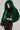 A person models a bold fashion outfit: the LUCKY HOODED FUR BOMBER in green, featuring the hood up, paired with a fitted black dress embellished with shiny details. The neutral background accentuates the vibrant and textured clothing.
