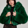 A person wearing the LUCKY HOODED FUR BOMBER in a vibrant green hue over a sheer black top with textured patterns stands against a plain background. They have long dark hair and wear large, dangling earrings.